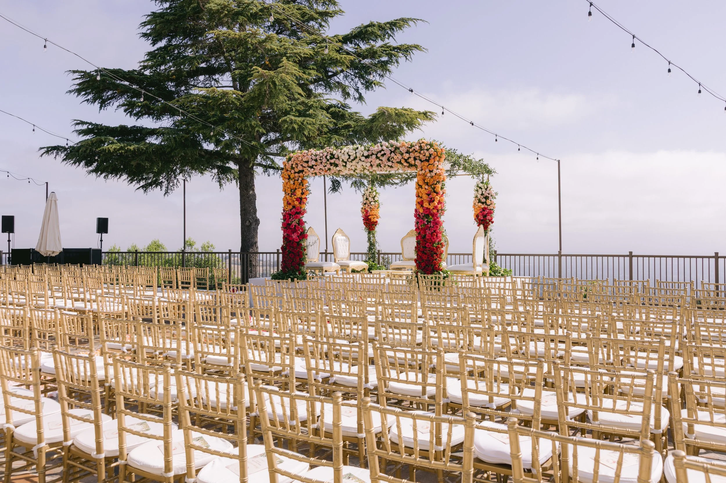 An outdoor wedding setup with rows of beige chairs facing a floral wedding arch, under a large tree with string lights overhead, overlooking a scenic view.