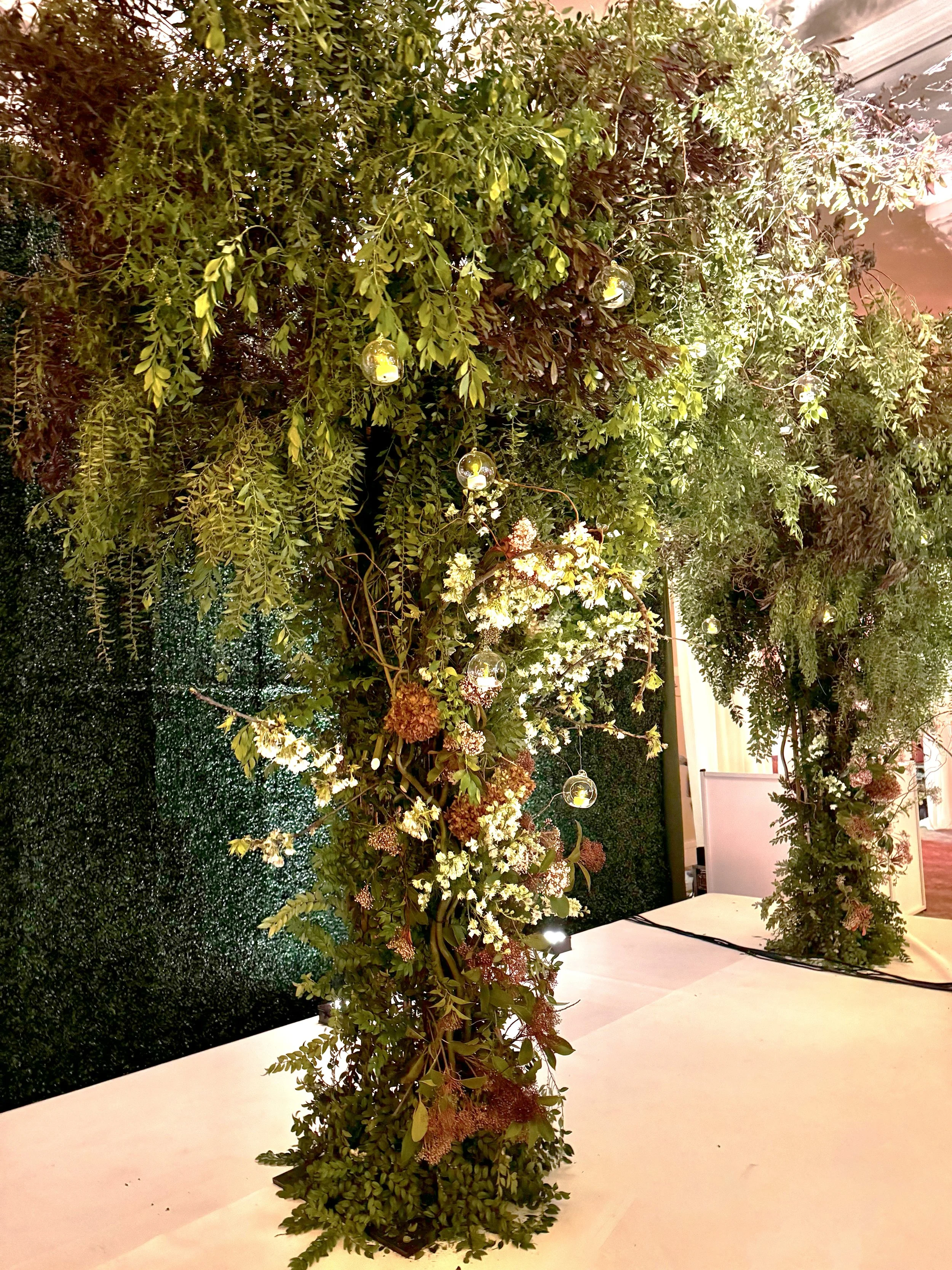 A vertical display of lush green foliage, hanging branches, and decorative glass globe lights, arranged as an indoor or outdoor decorative installation against a textured wall.
