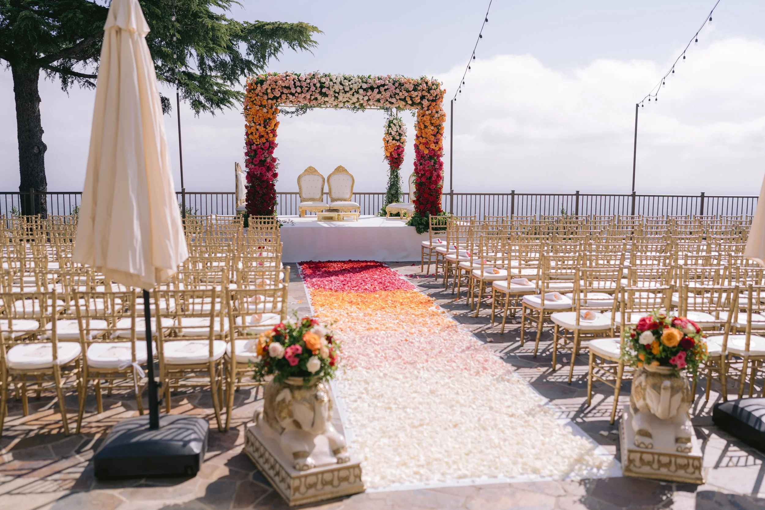 Outdoor wedding ceremony setup with a flower arch, chairs arranged on either side of a flower petals aisle, and two ornate chairs at the altar, under string lights, with a view of the sky and distant horizon.