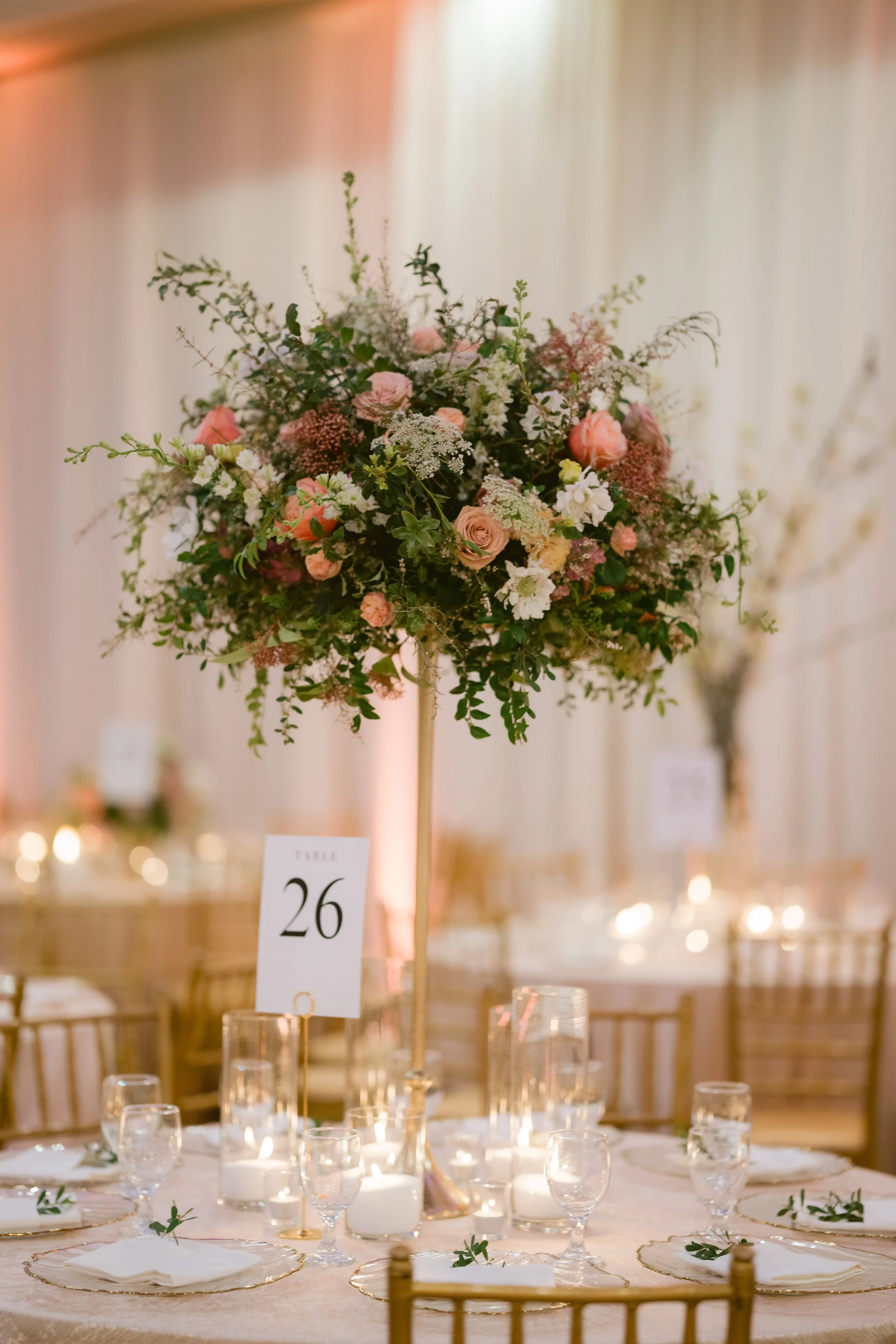 Elegant banquet table decorated with a large floral centerpiece consisting of pink, white, and cream flowers and lush greenery. The table is set with white plates, gold-rimmed glasses, white napkins, and small greenery accents. A small table number s
