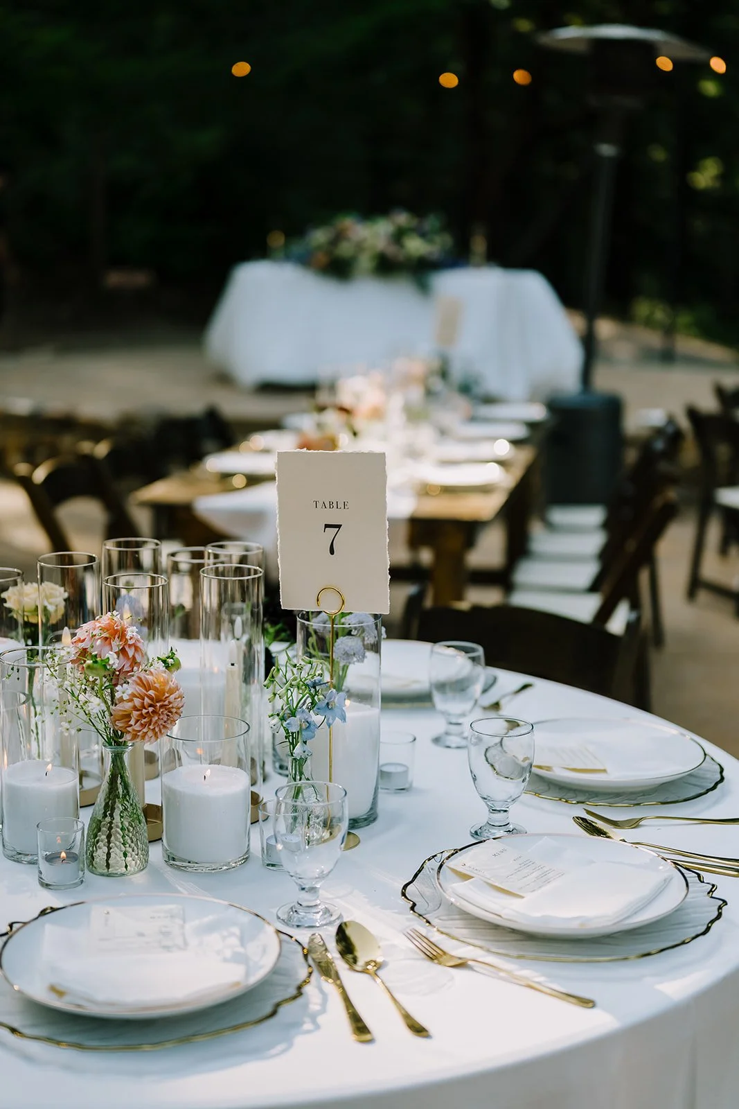 Elegant outdoor wedding table setting with white plates, gold utensils, glassware, and floral centerpieces, with a table number sign indicating 'Table 7' in the foreground and a blurred background of additional tables and greenery.