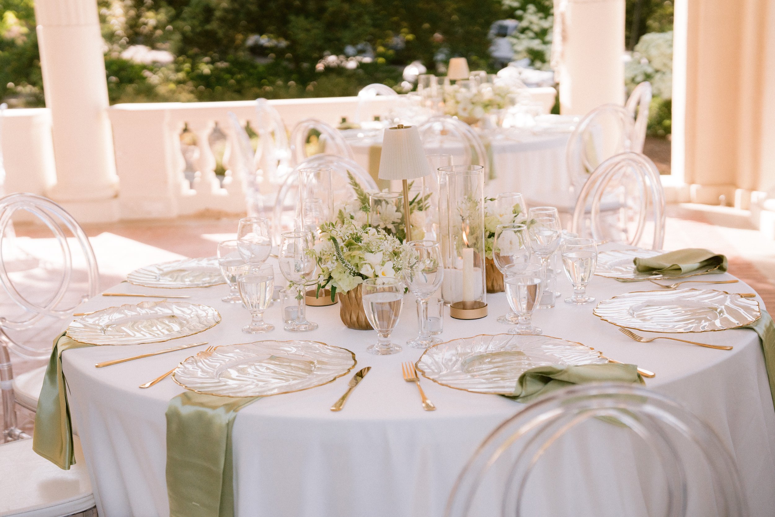 Elegant outdoor banquet table with white tablecloth, floral centerpiece, candles, glassware, and gold-rimmed plates, set on a porch with columns and greenery in the background.
