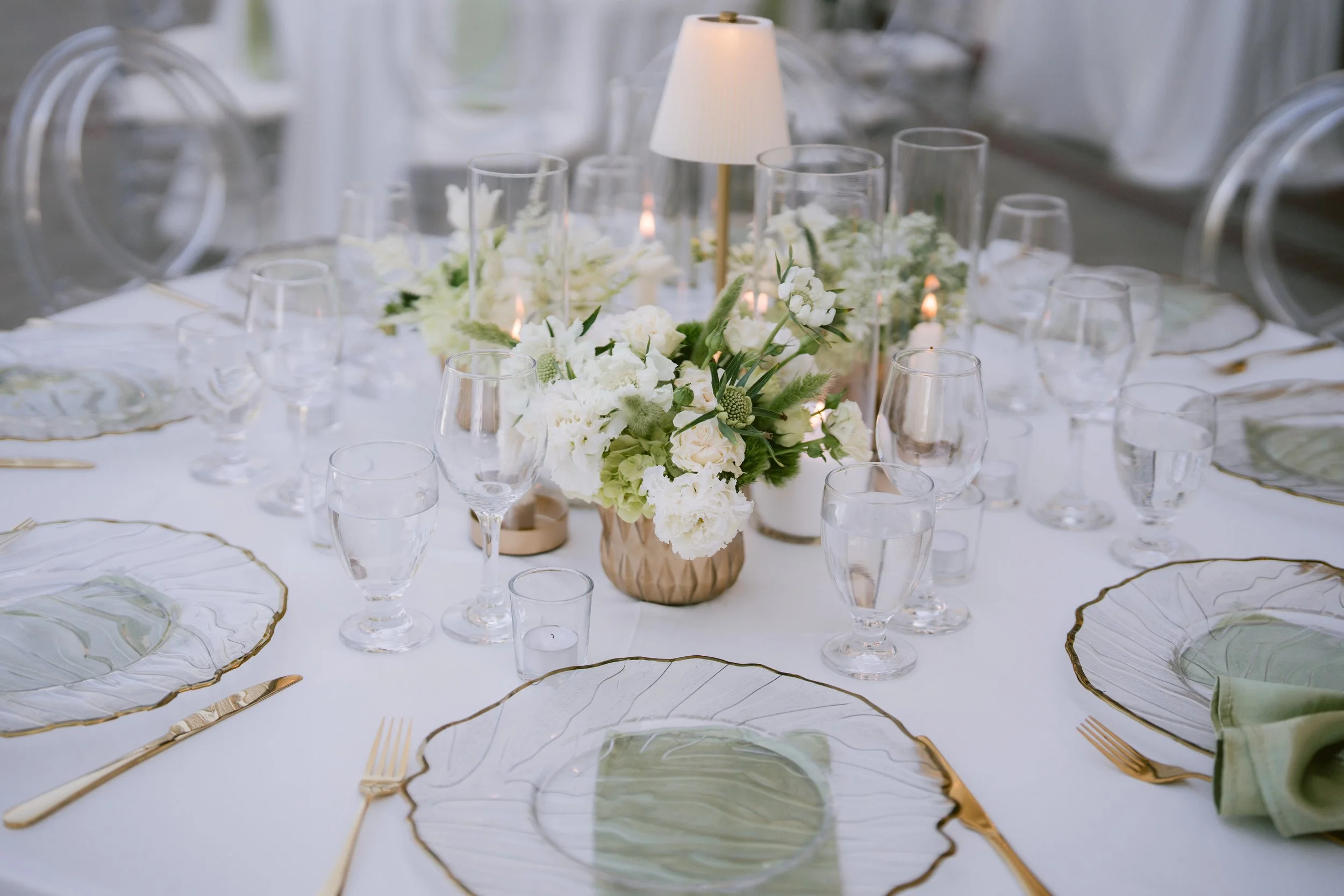 Elegant dining table decorated with a floral centerpiece of white flowers and greenery, surrounded by clear glassware, gold utensils, and unique leaf-shaped plates with gold edges. A small table lamp provides soft lighting.