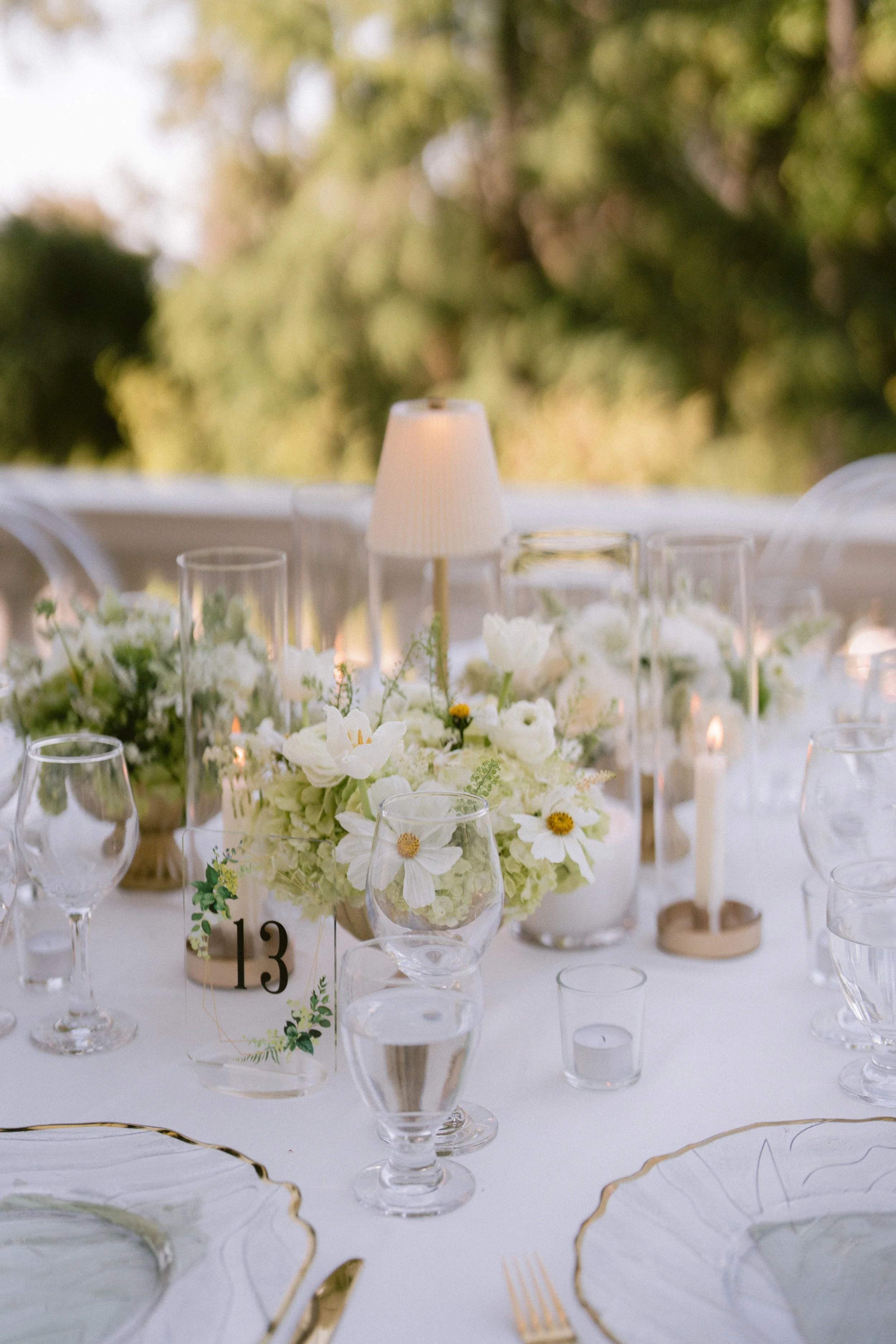 Elegant table setting with a floral centerpiece, multiple clear glassware, small candles, and a white tablecloth, outdoors with a blurred background of trees and greenery.