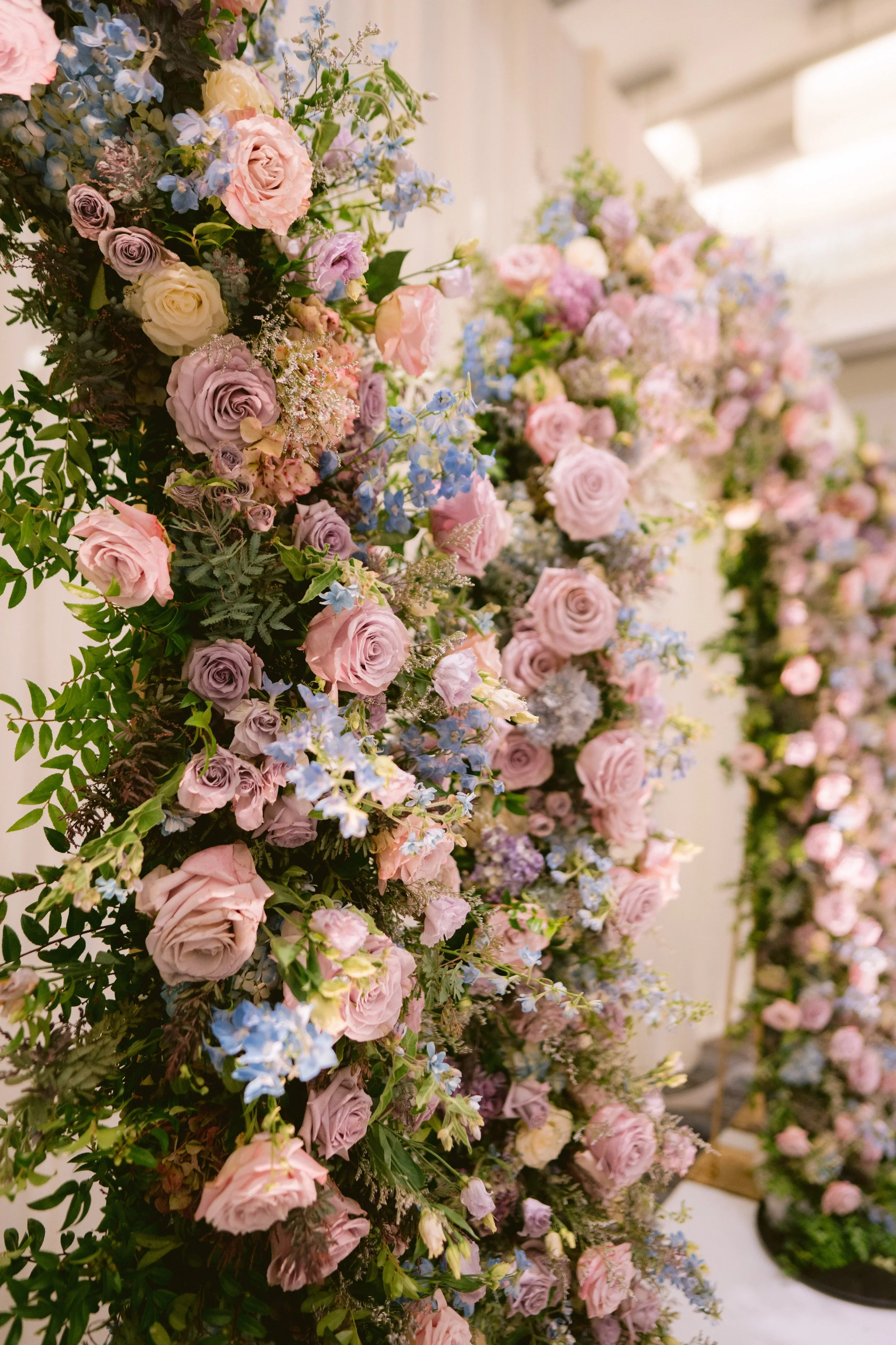 Close-up of a cascading floral arrangement featuring pink roses, purple roses, blue flowers, and green foliage in a decorative setting.