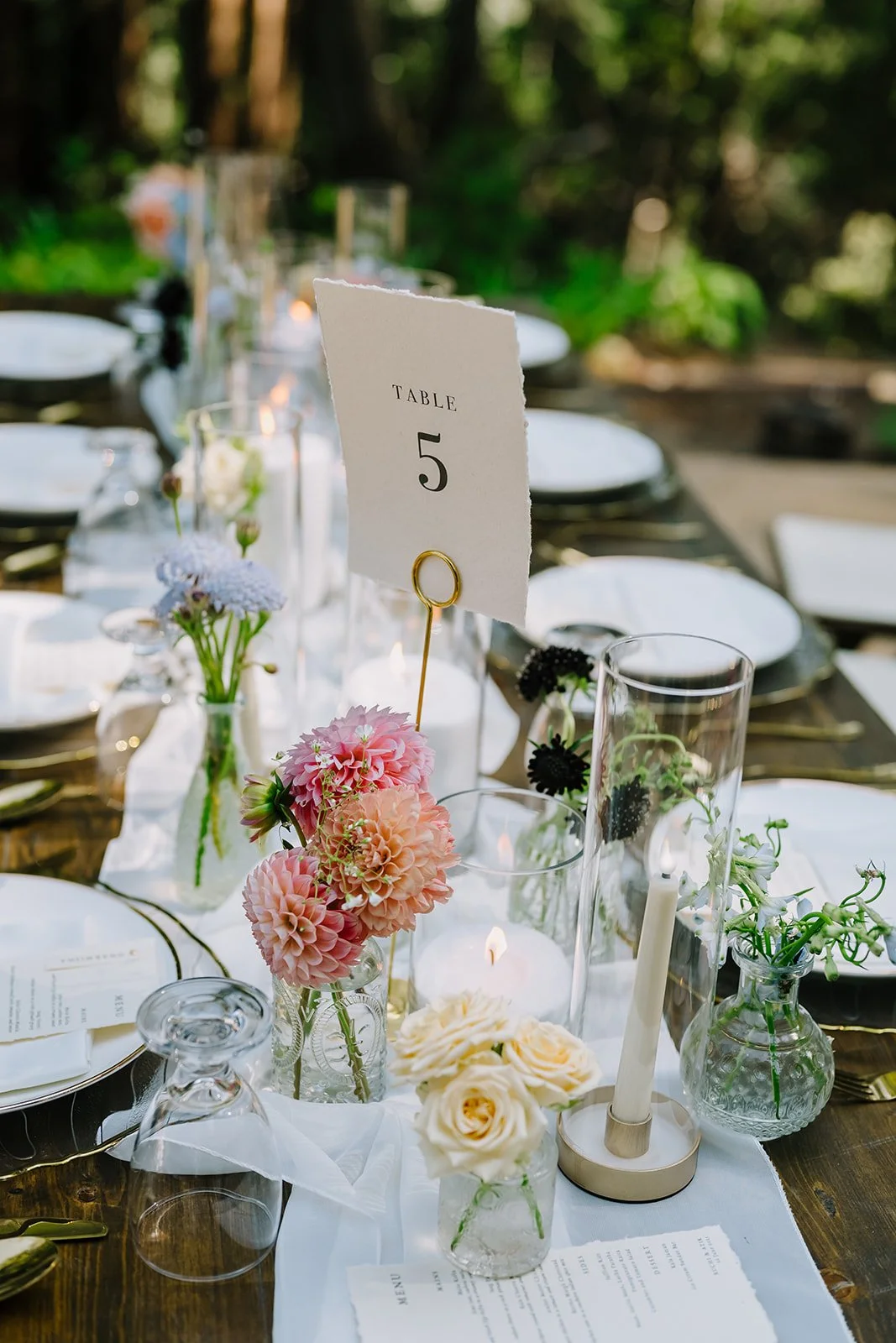 A beautifully set outdoor dining table with floral centerpieces, candles, glasses, and table number 5 card in a garden setting.