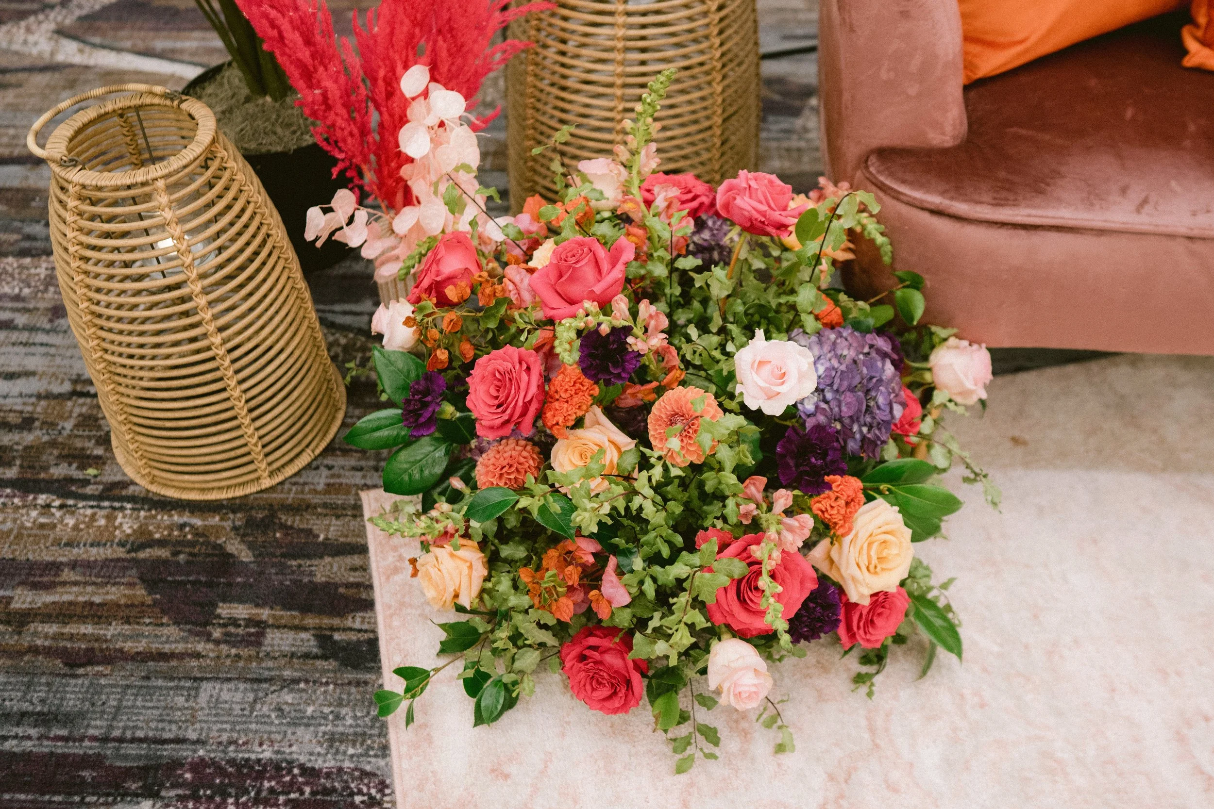 Colorful flower arrangement with pink, purple, orange, and peach flowers next to wicker lanterns and a pink sofa on a rustic wooden floor.