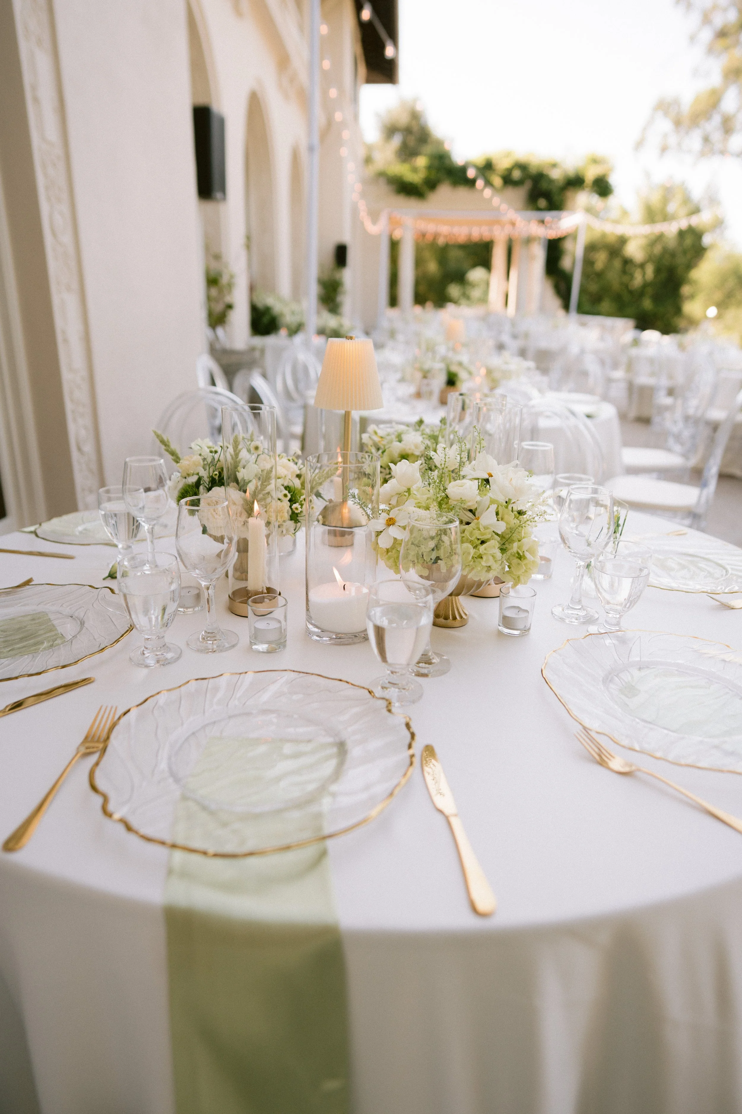Elegant outdoor wedding reception table with floral centerpieces, candles, and glassware, set against a backdrop of trees and string lights.