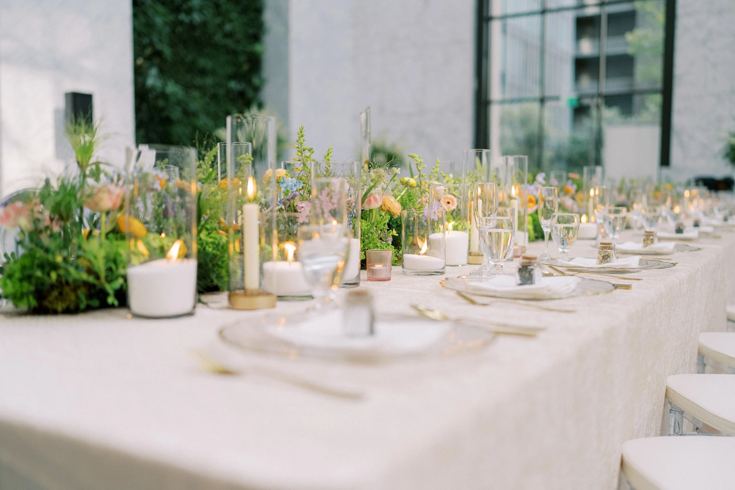 Elegant outdoor event table decorated with candles and lush floral arrangements, set against a modern building and greenery.