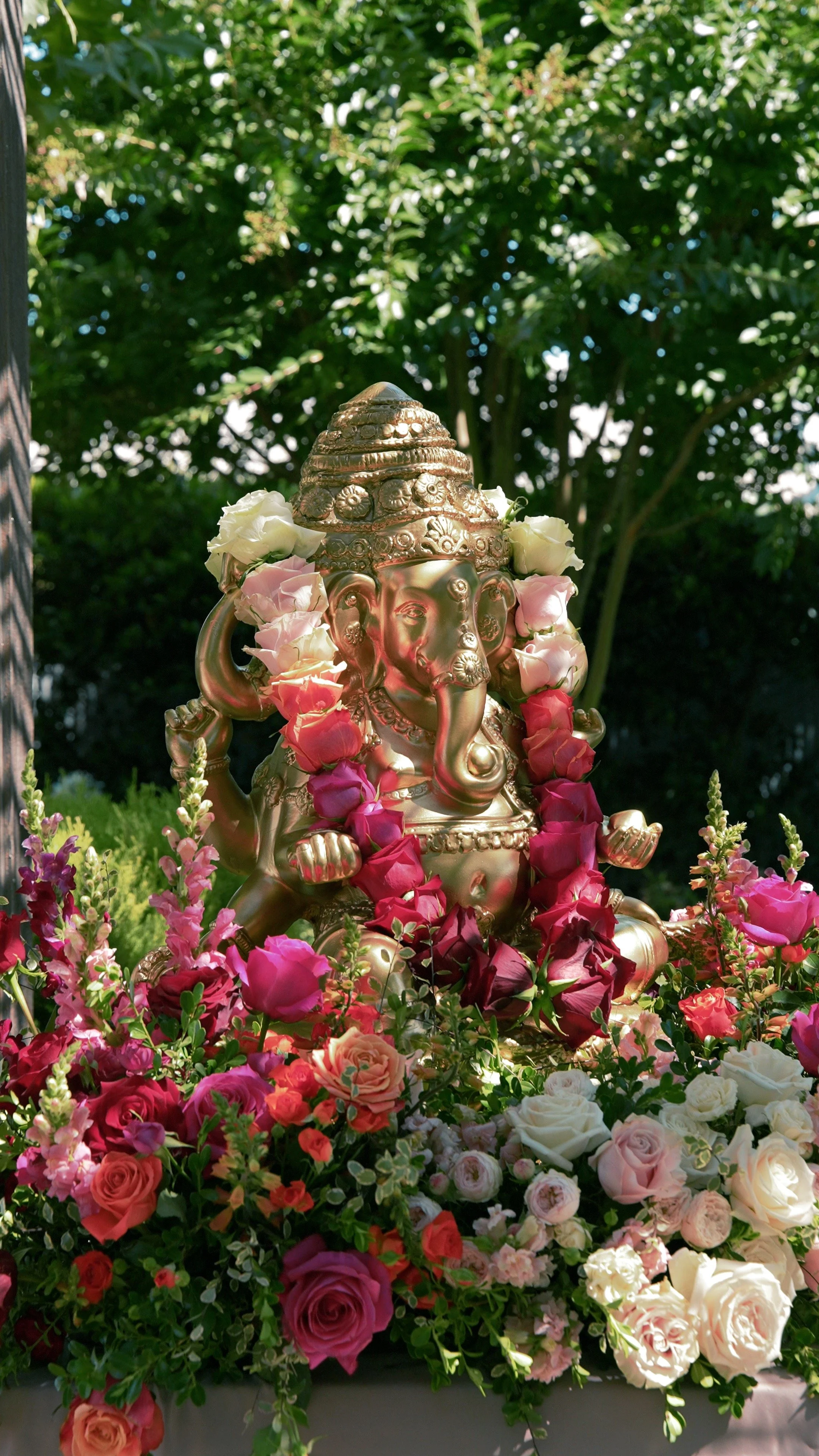 A golden statue of the Hindu deity Ganesha surrounded by pink, white, and red flowers outdoors with green foliage in the background.