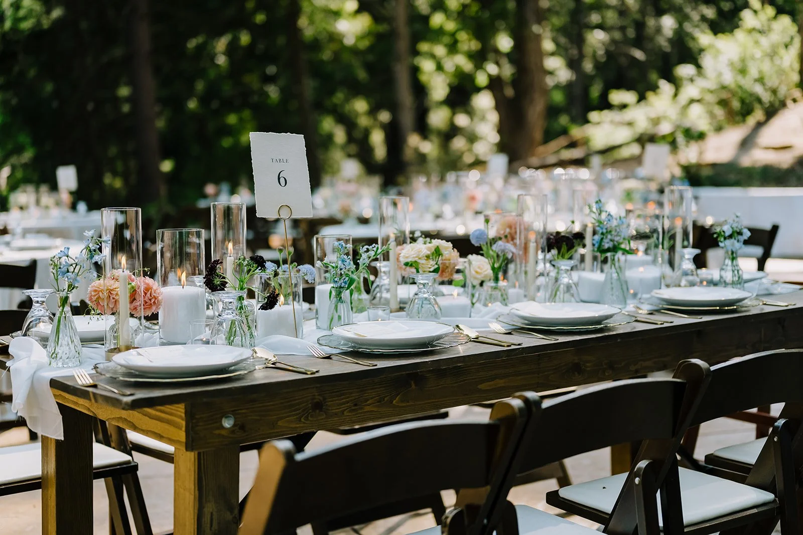 Outdoor wedding reception table decorated with candles, flowers, and tableware, surrounded by trees.