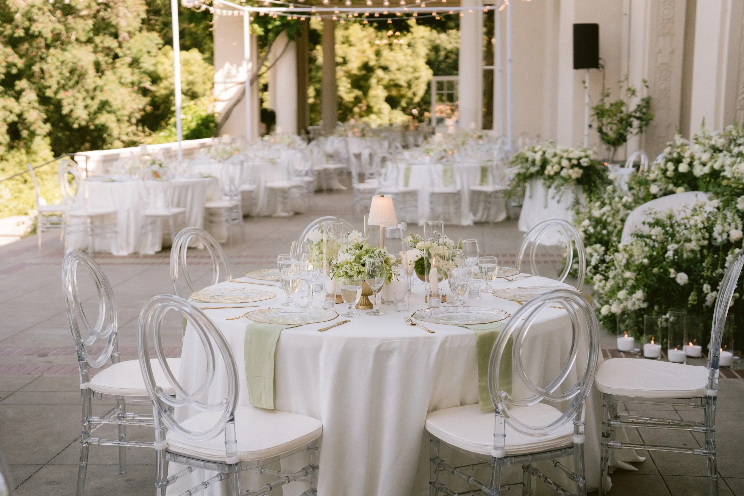 Elegant outdoor event setup with a round table covered in a white tablecloth, decorated with floral centerpieces, candles, and glassware. Clear ghost chairs surround the table, and additional tables and white floral arrangements are visible in the ba