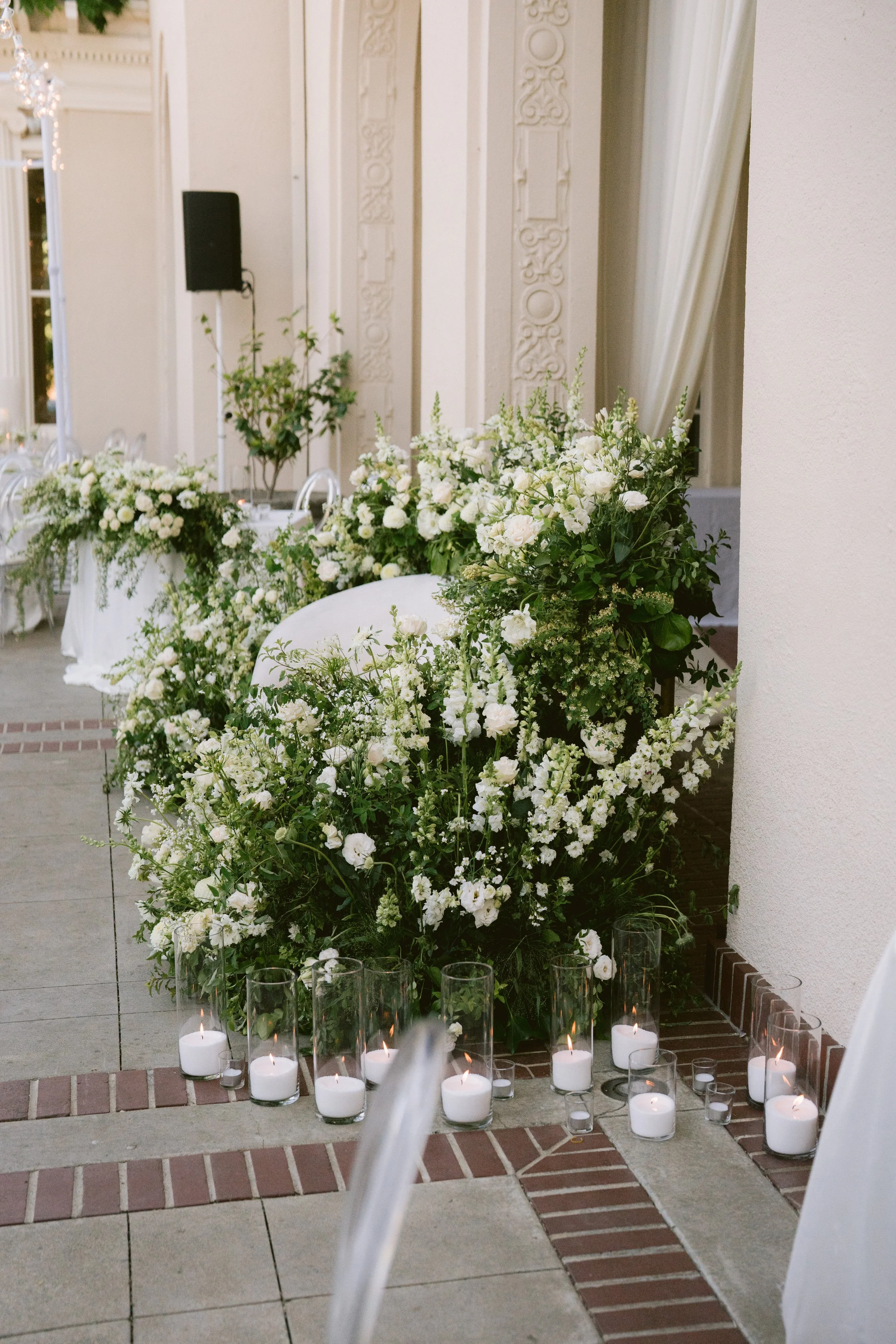 A floral arrangement of white flowers and greenery on a table at an elegant indoor event, with candles in clear glass holders placed on the floor in front of the flowers.