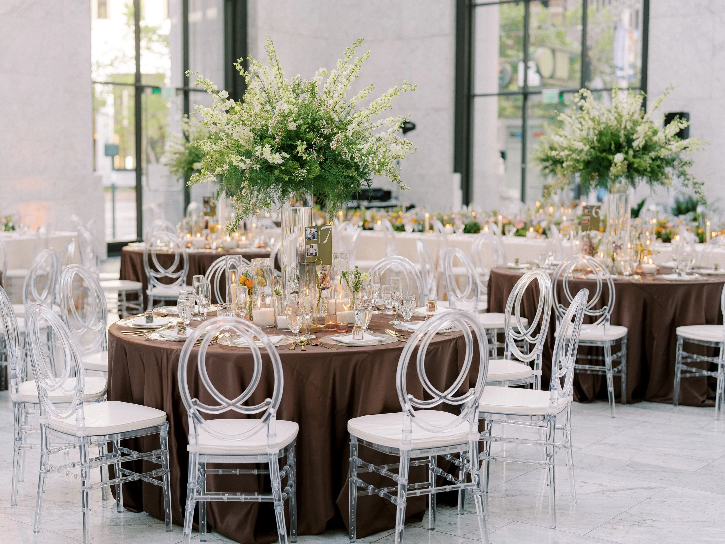 Elegant banquet event in a modern venue with large floral centerpieces featuring white flowers on round tables with brown tablecloths and clear acrylic chairs.