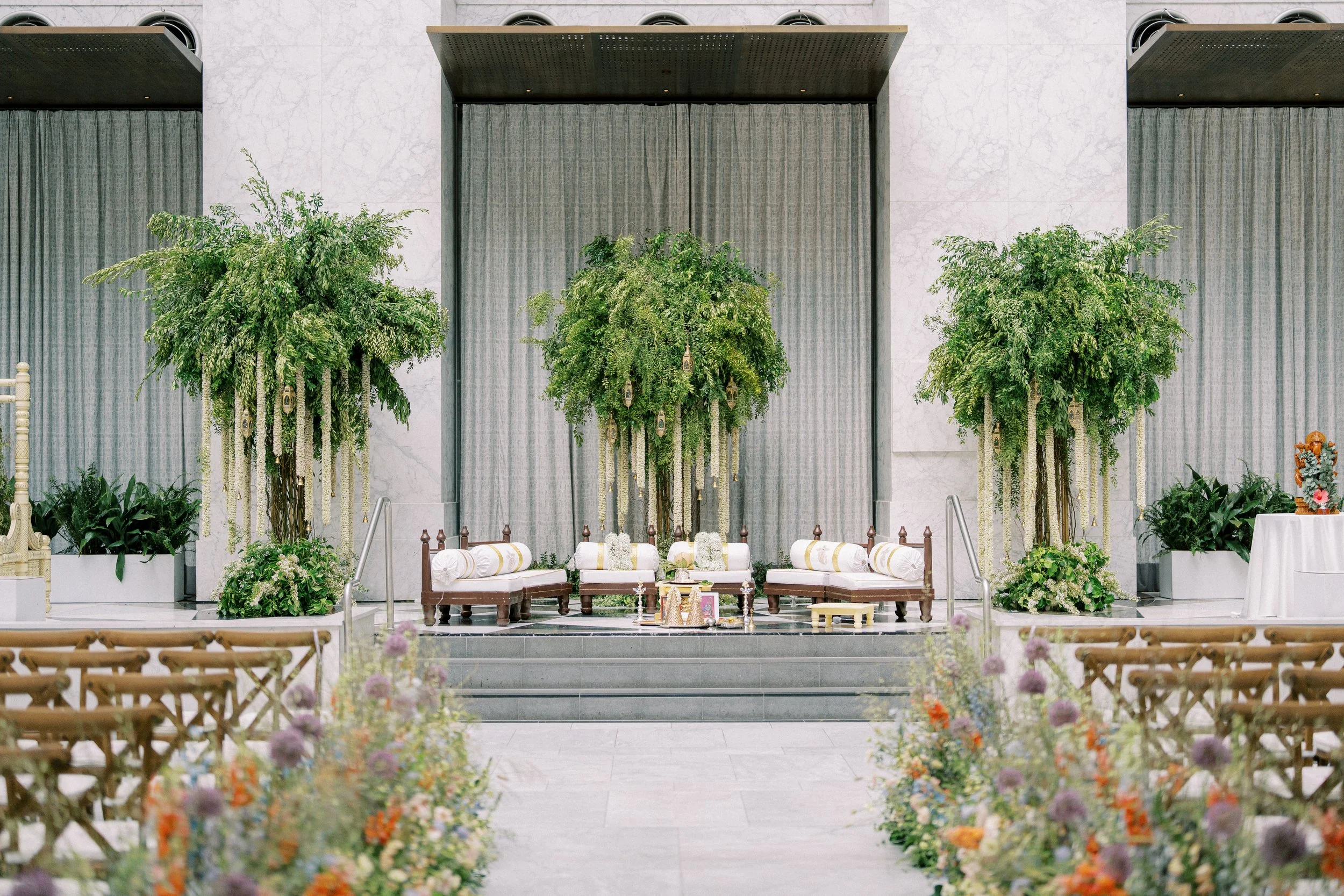 A decorated stage with green plants and floral arrangements, white cushioned chairs, and a table, set up for a ceremony or event in an indoor venue with marble walls and curtains.