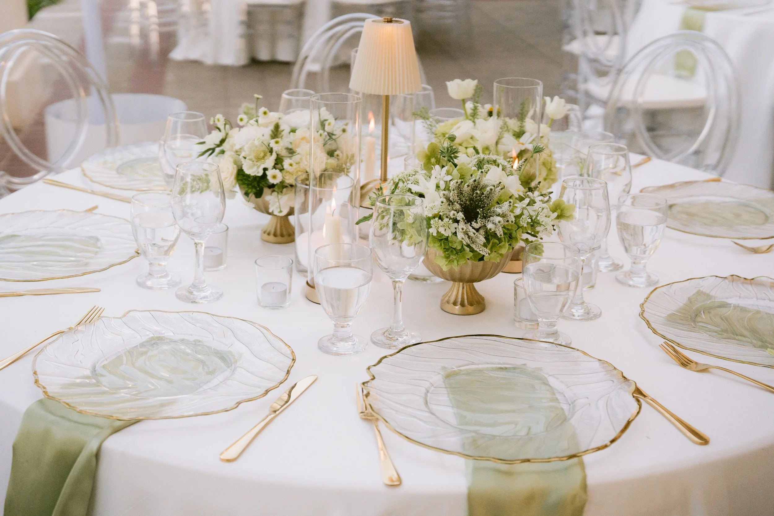 Elegant dining table decorated with white and green floral arrangements, glassware, and gold-accented plates and utensils, with a small table lamp in the center.