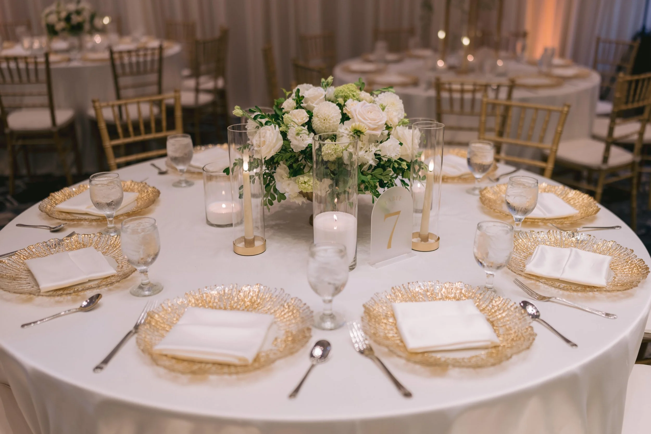 A round banquet table decorated with a large floral centerpiece of white roses and greenery. The table has four glasses of water, gold charger plates with white napkins, and gold-rimmed candleholders with white candles. The background shows multiple 