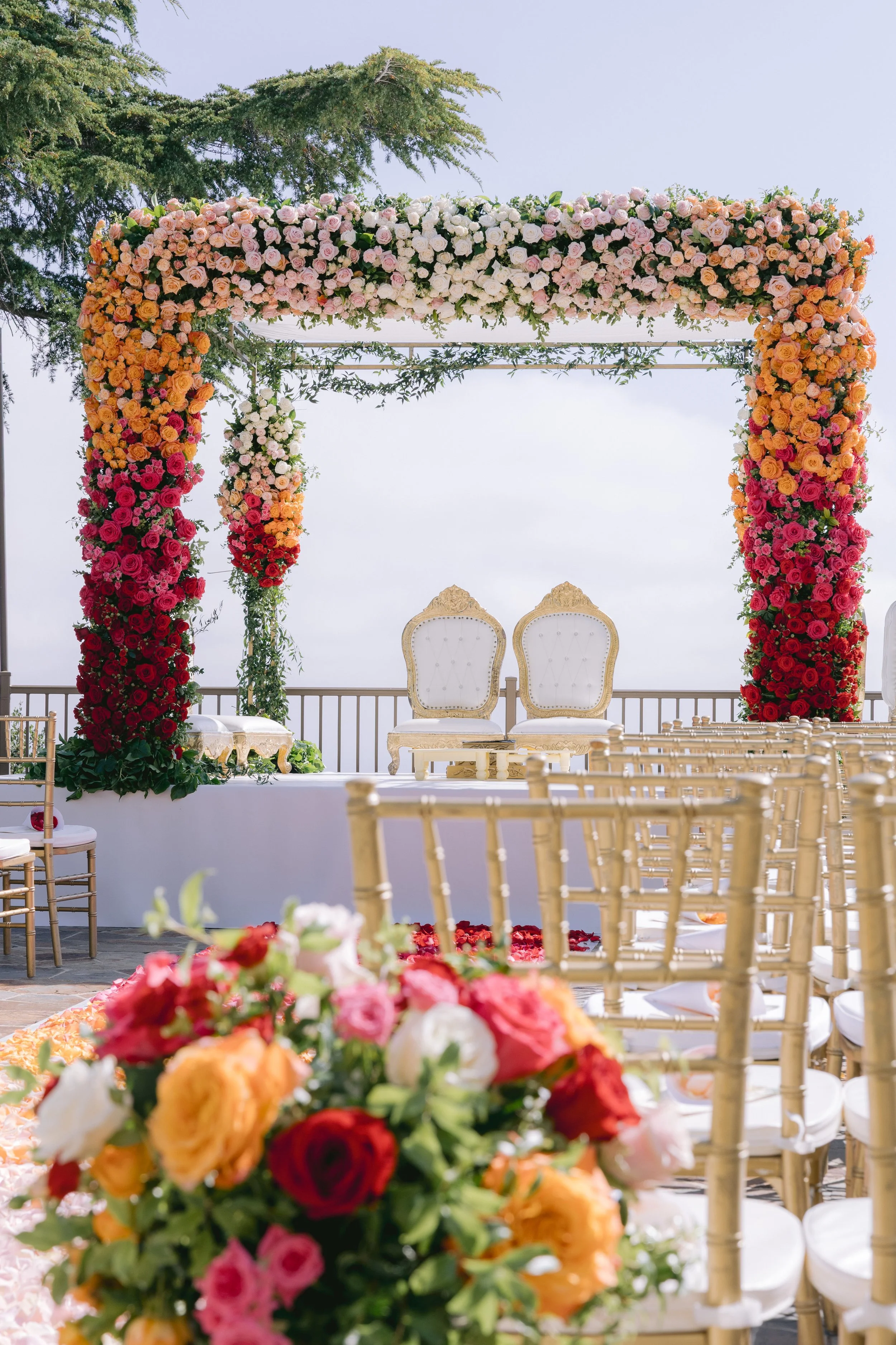 Flower-covered wedding arch with white chairs in front, set outdoors on a terrace with a clear sky and trees in the background.
