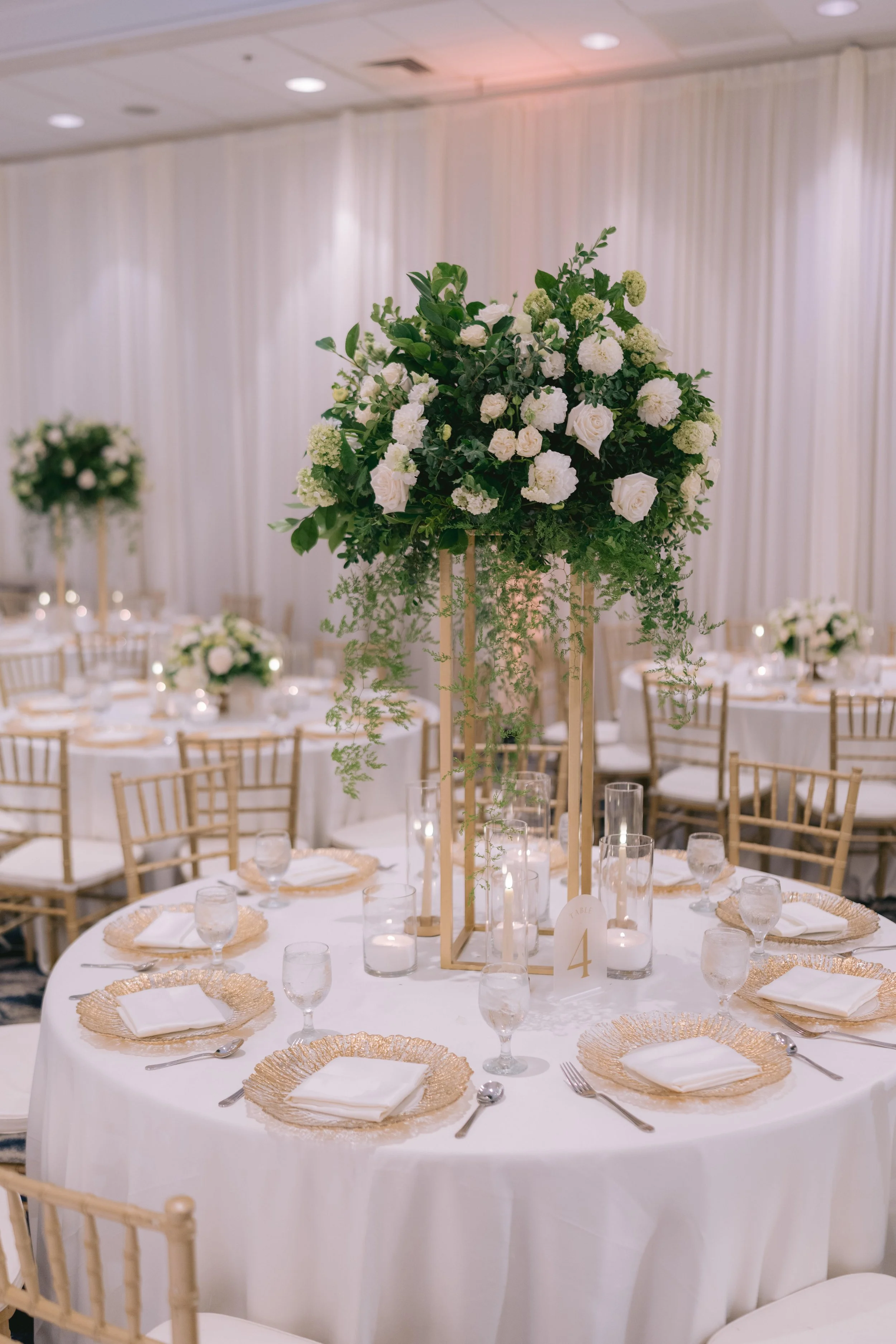 Elegant round banquet table set for a formal event with gold-rimmed plates, white napkins, water glasses, and gold chairs, with a tall floral centerpiece of white roses and greenery in a wooden frame, in a decorated banquet hall.