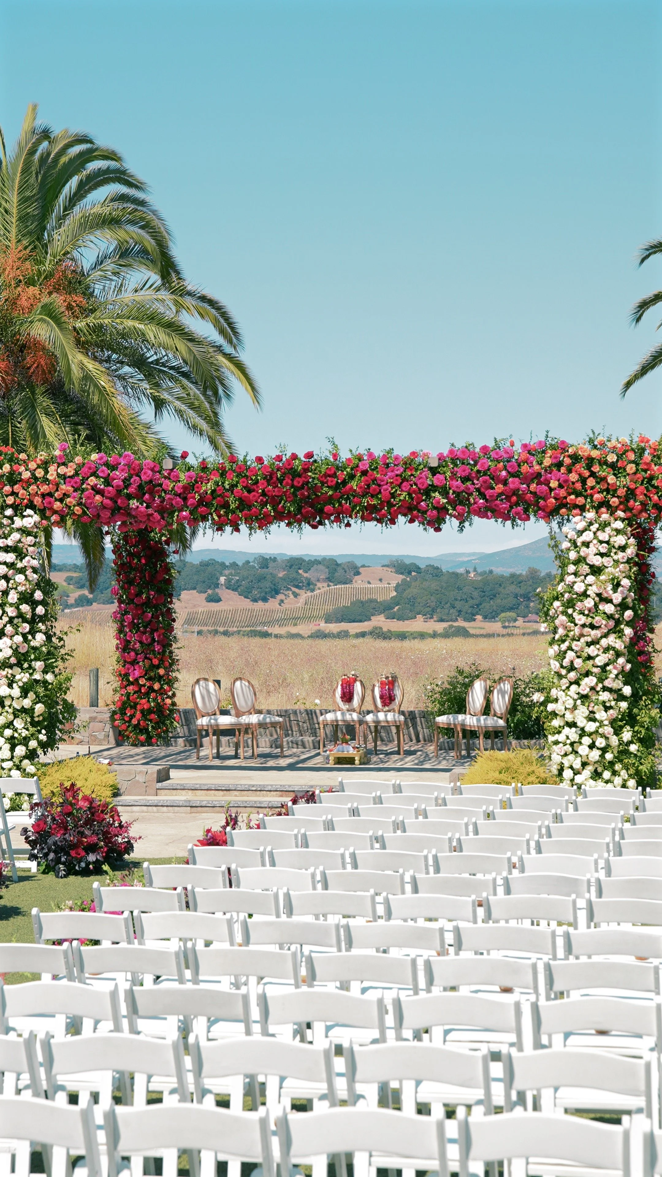 Outdoor wedding ceremony setup with white chairs, a floral archway, and a backdrop of rolling hills under a clear blue sky.