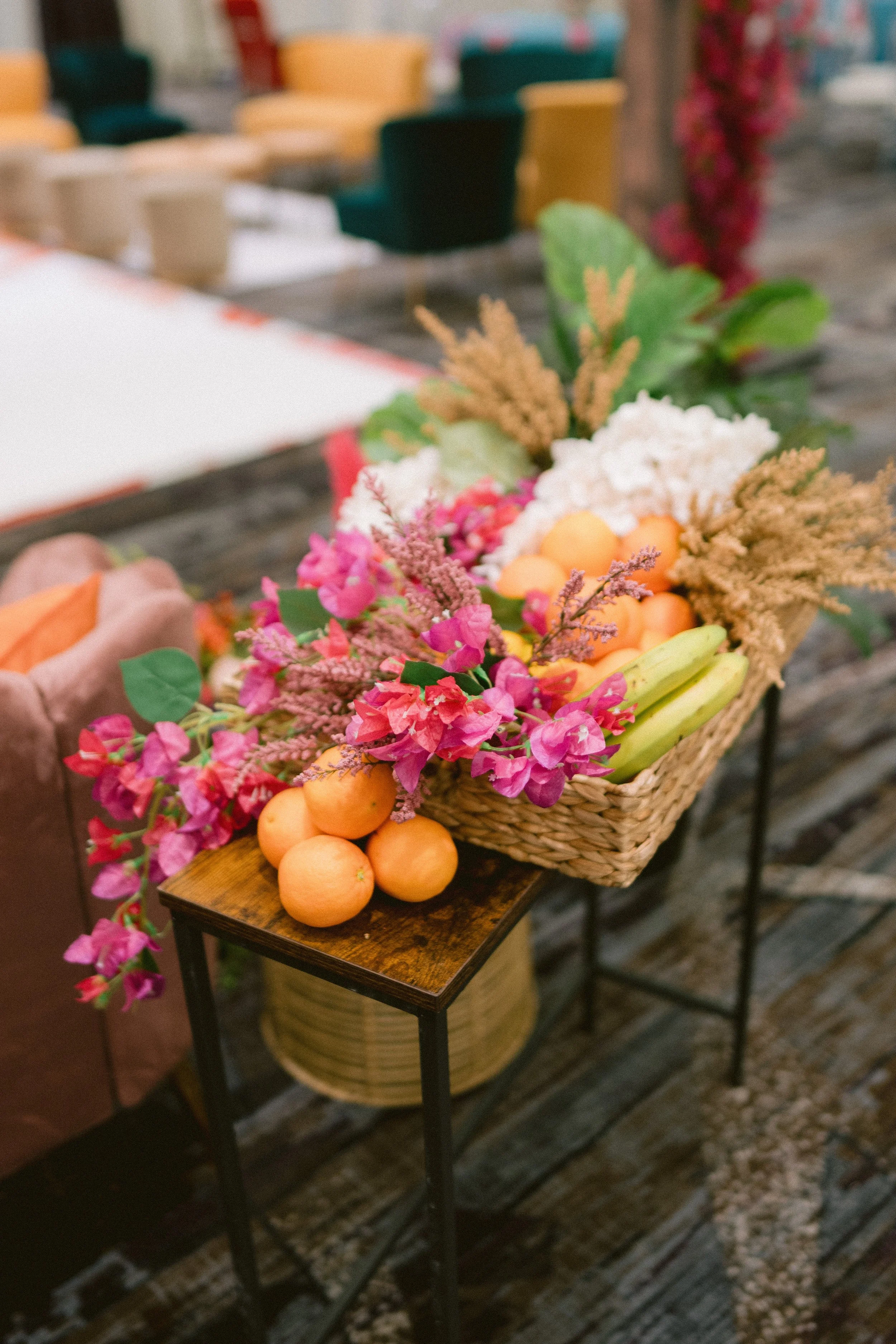 A basket of pink, white, and orange flowers along with mangoes on a small wooden table, with a background of multicolored chairs and a stove.