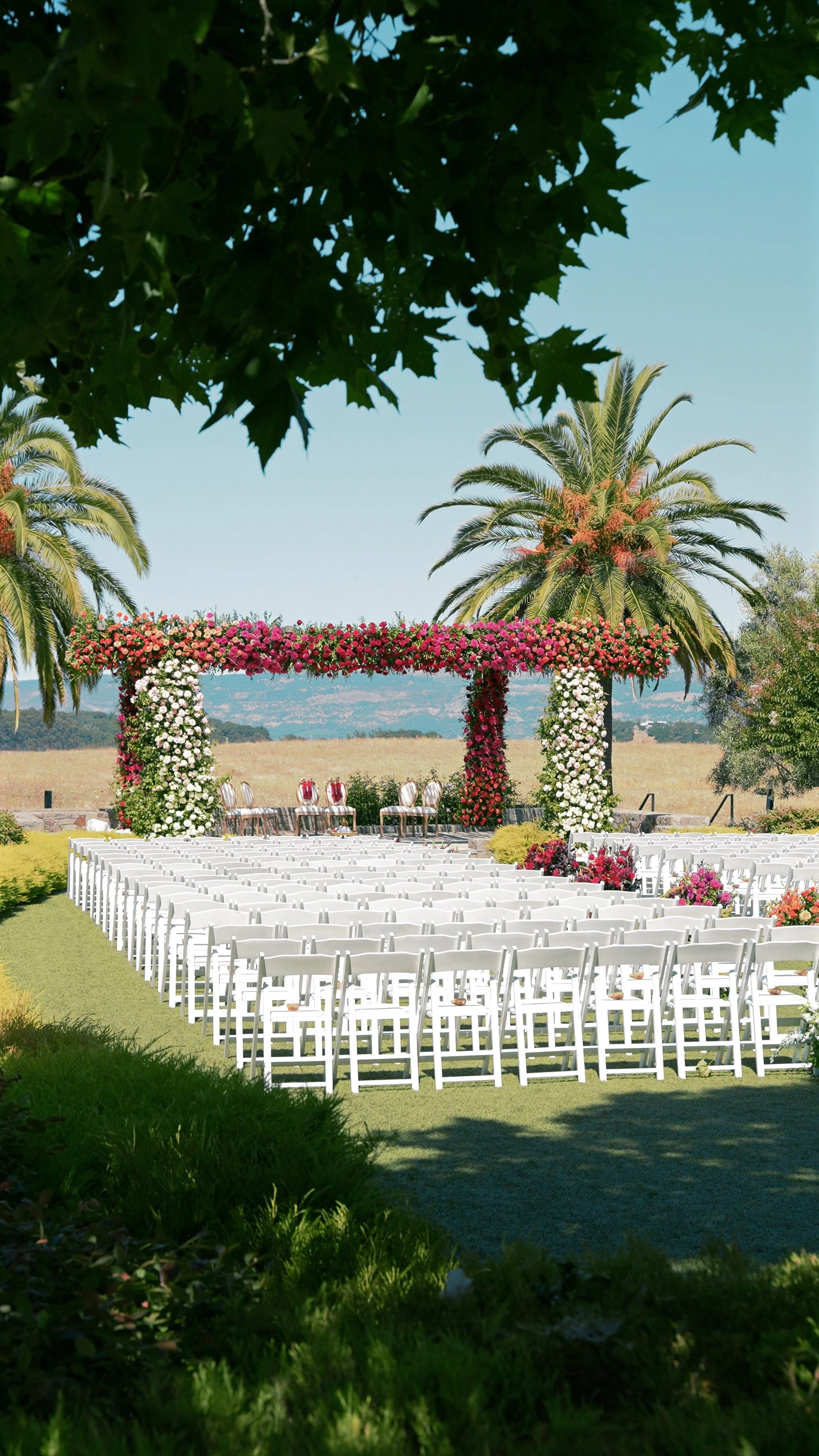Outdoor wedding ceremony setup with white chairs, floral arch, and palm trees in the background.
