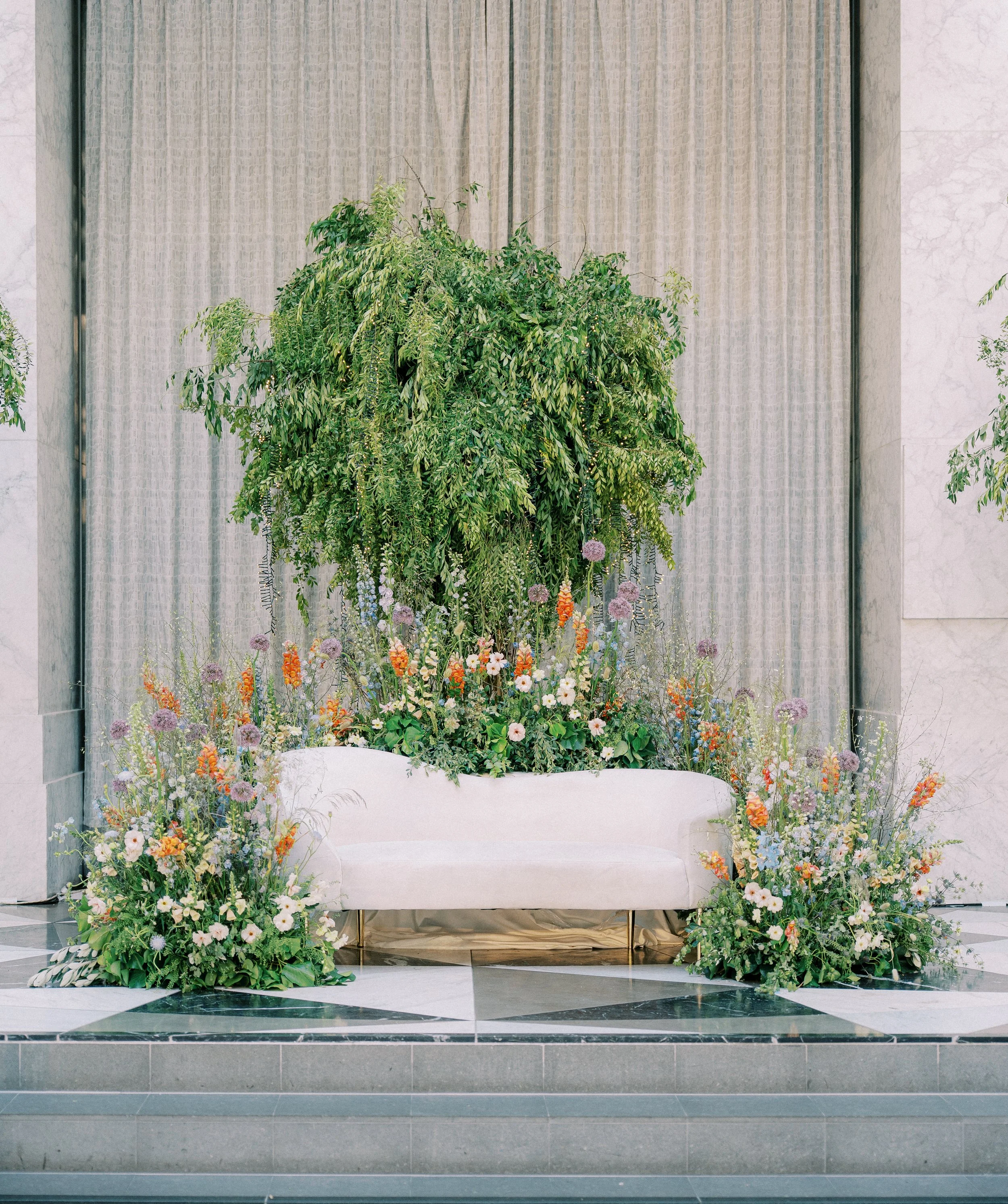A floral arrangement with a large green leafy plant and a variety of colorful flowers is set up in front of a beige curtain. The display is on a marble floor in an indoor setting.