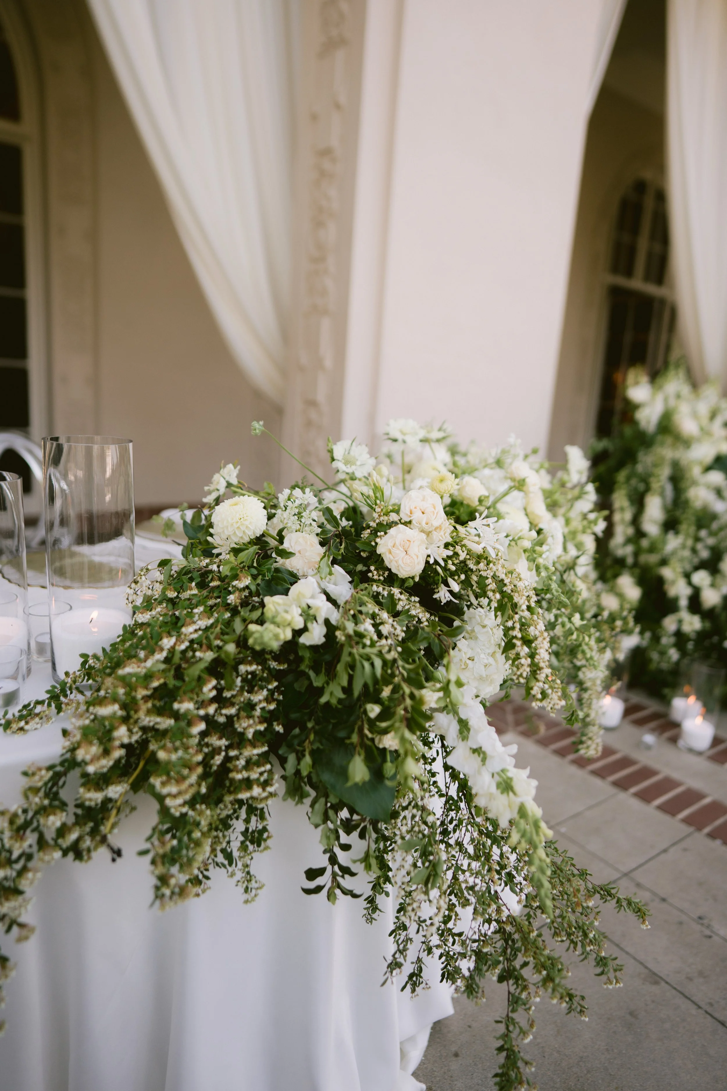 A floral arrangement of white flowers and greenery on a table, with a white draped backdrop and candles on the floor nearby.
