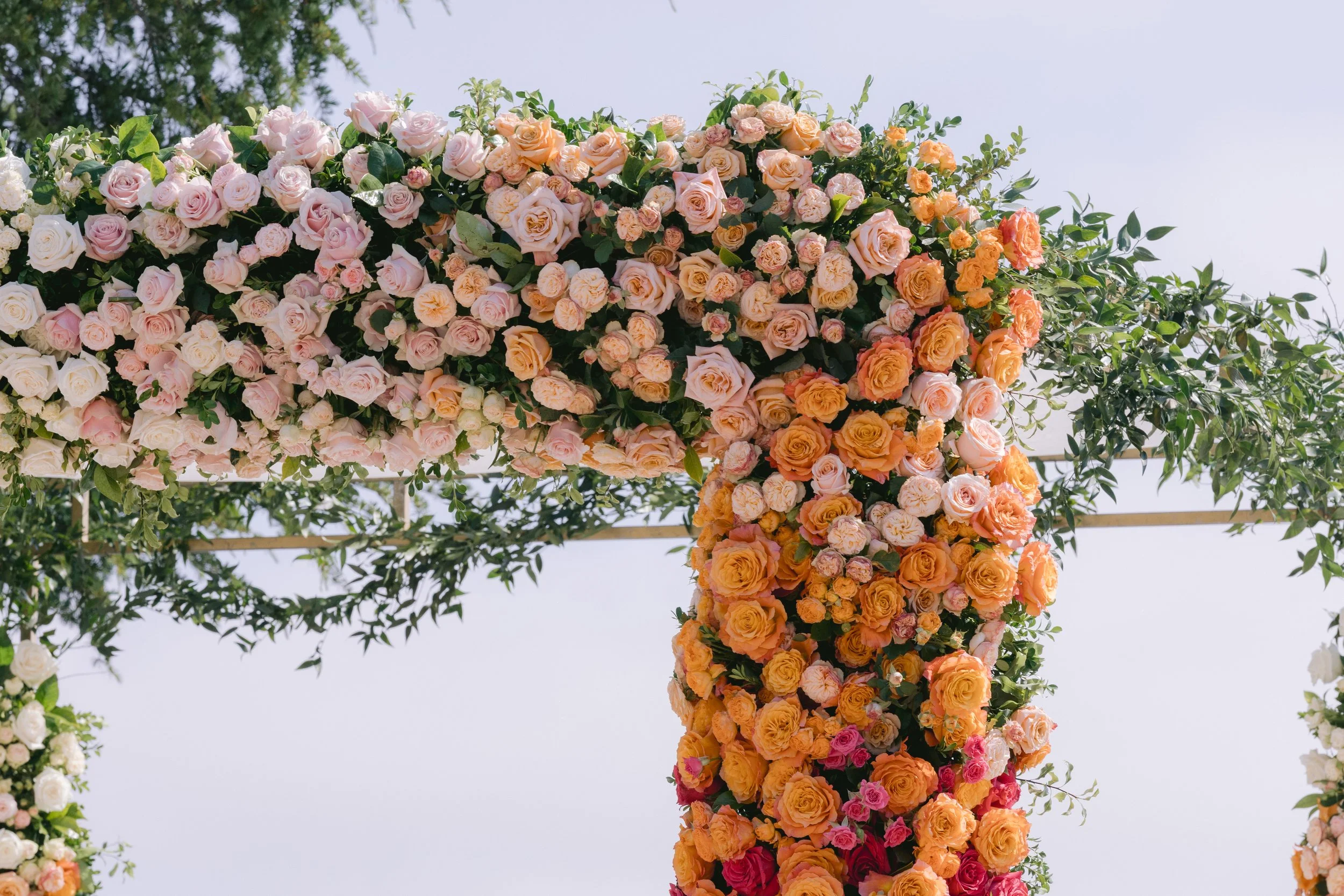 A large floral arrangement of pink, yellow, orange, and white roses decorated with green leaves, set against a clear sky background.