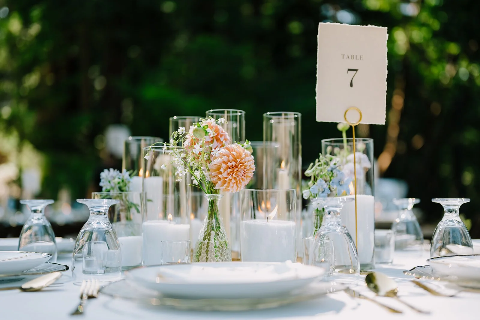 Elegant outdoor table setting with floral centerpieces in glass vases, surrounded by candles and upside-down wine glasses, with a table number sign reading 'Table 7' on a gold stand, set against a lush green background.