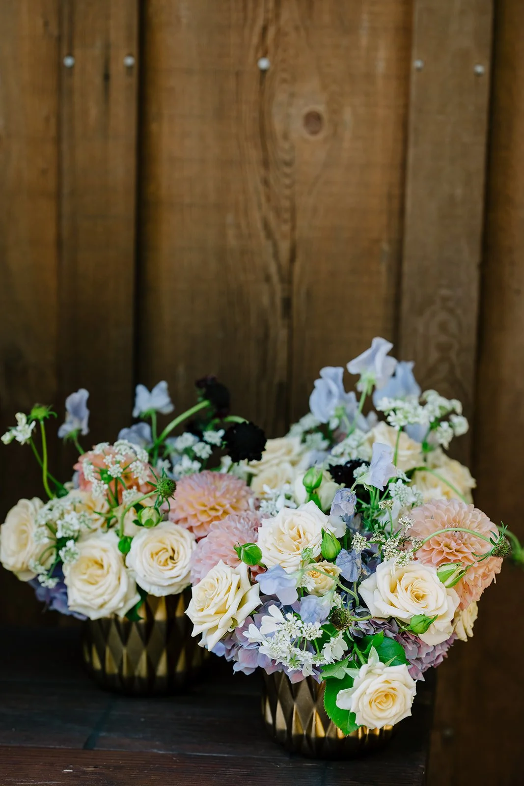 Two decorative flower arrangements in gold vases with white, pink, purple, and black flowers set against a wooden wall.