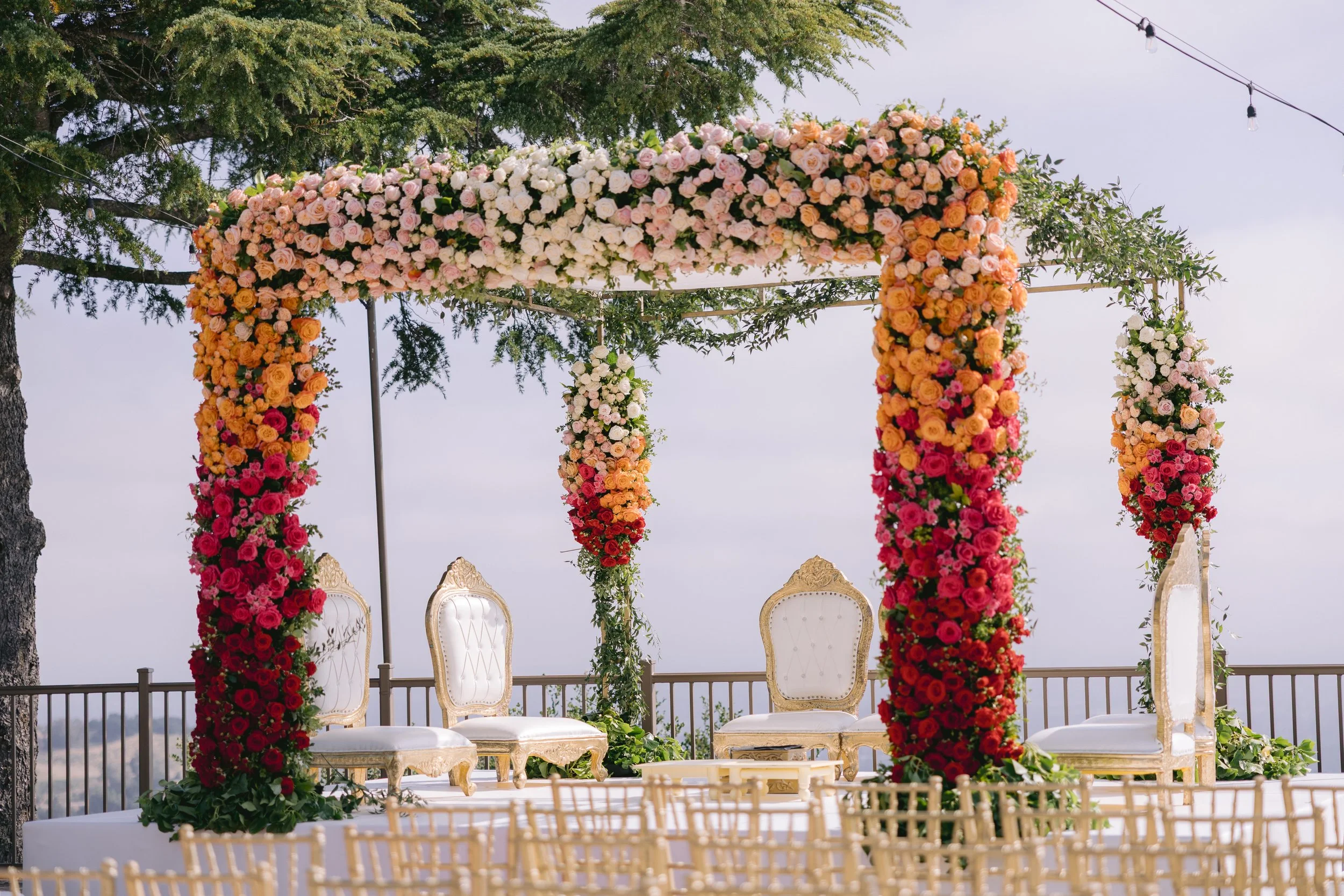 A decorated outdoor wedding altar with a floral canopy of mostly pink, white, and orange roses, and white chairs arranged for the ceremony, under a string of lights, with trees and a cloudy sky in the background.