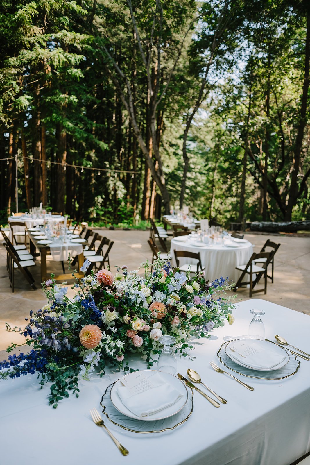 Wedding reception setup outdoors in a forest, with round and long tables decorated with floral arrangements, fine china, glassware, and gold cutlery.