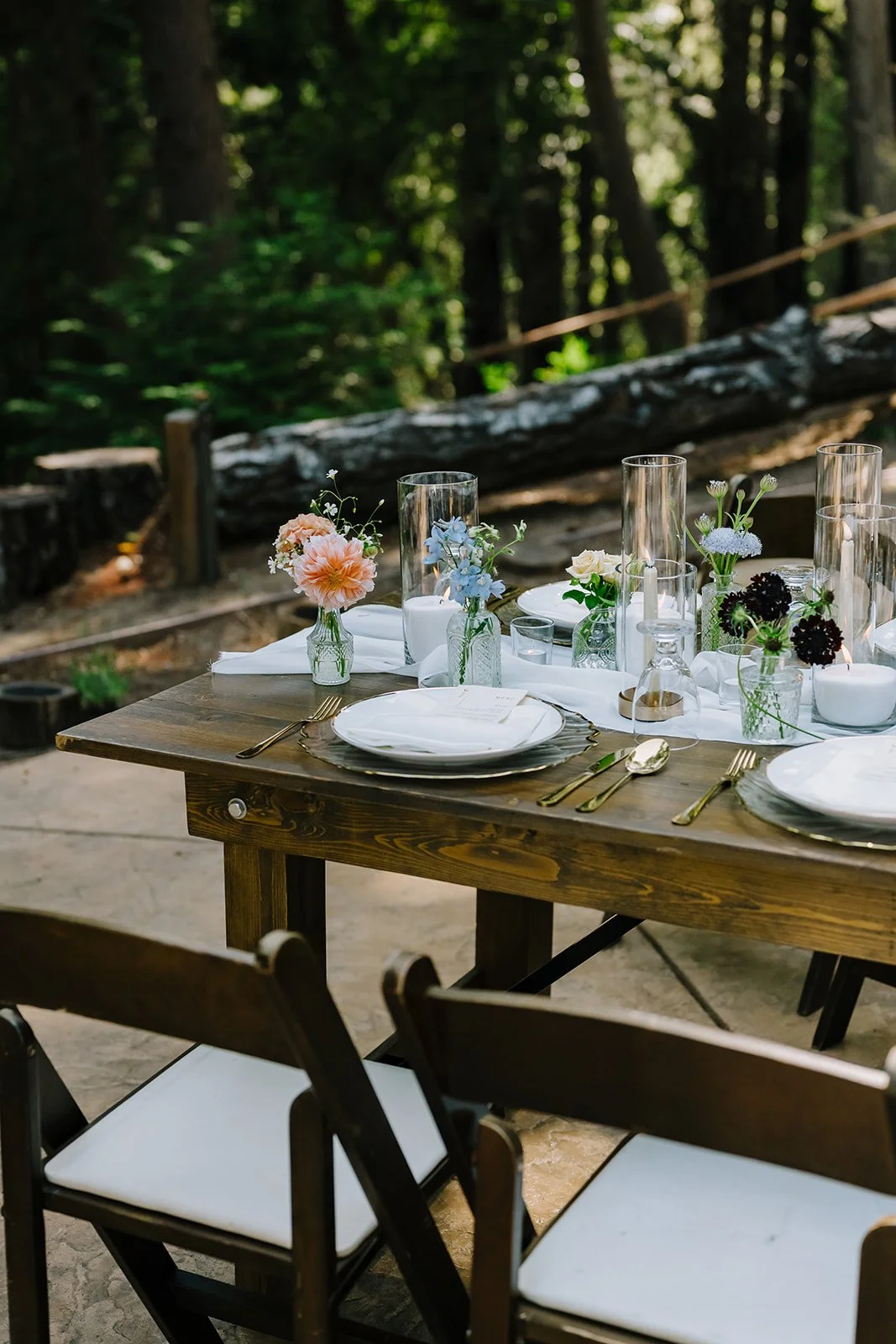 Outdoor table setting with white plates, gold cutlery, floral centerpieces in glass vases, and large glass candle holders on a rustic wooden table amidst a forest background.