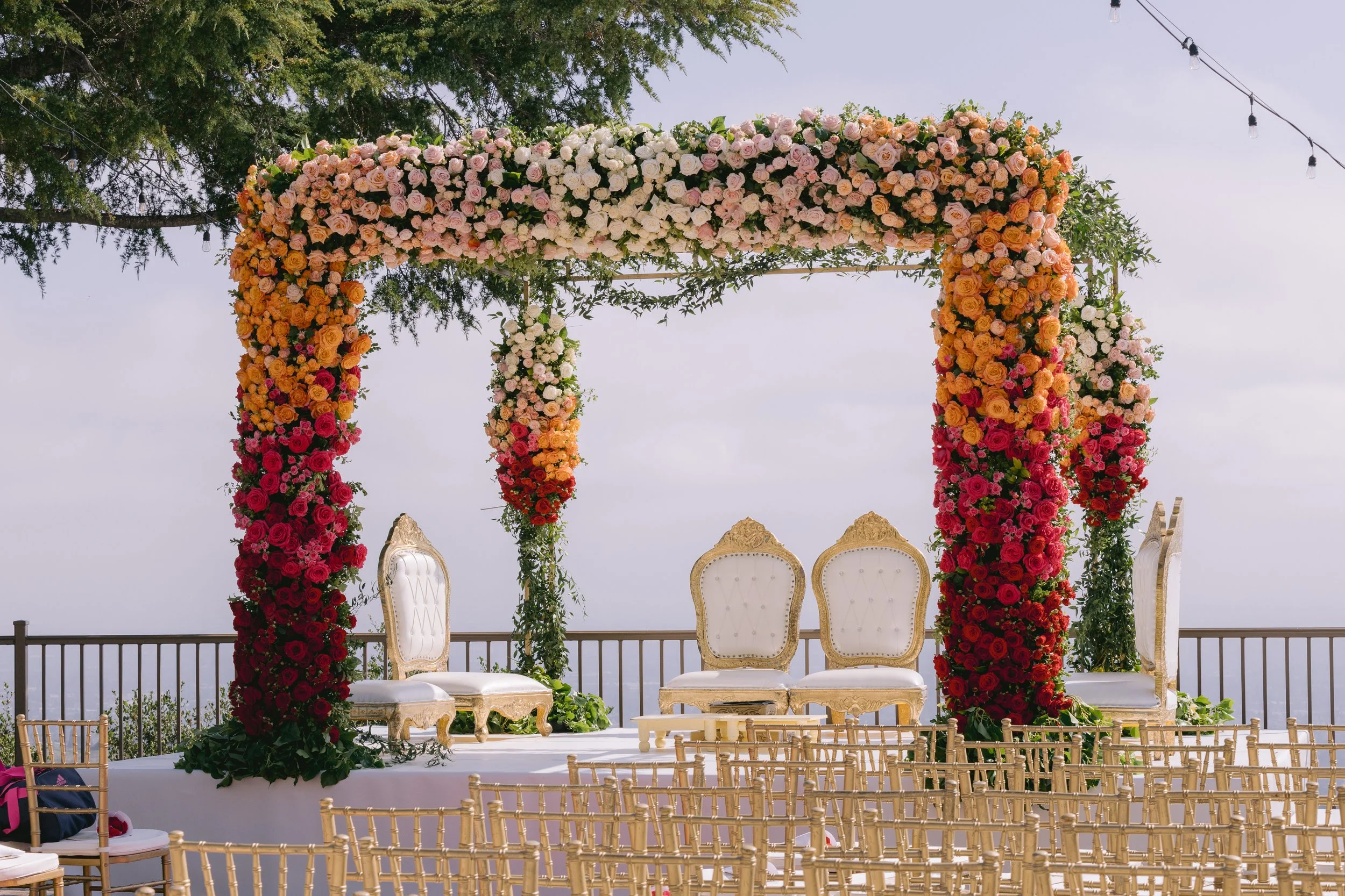 A wedding arch decorated with pink, white, orange, red, and peach flowers, set up outdoors on a terrace with chairs arranged for a ceremony, and string lights hanging overhead.