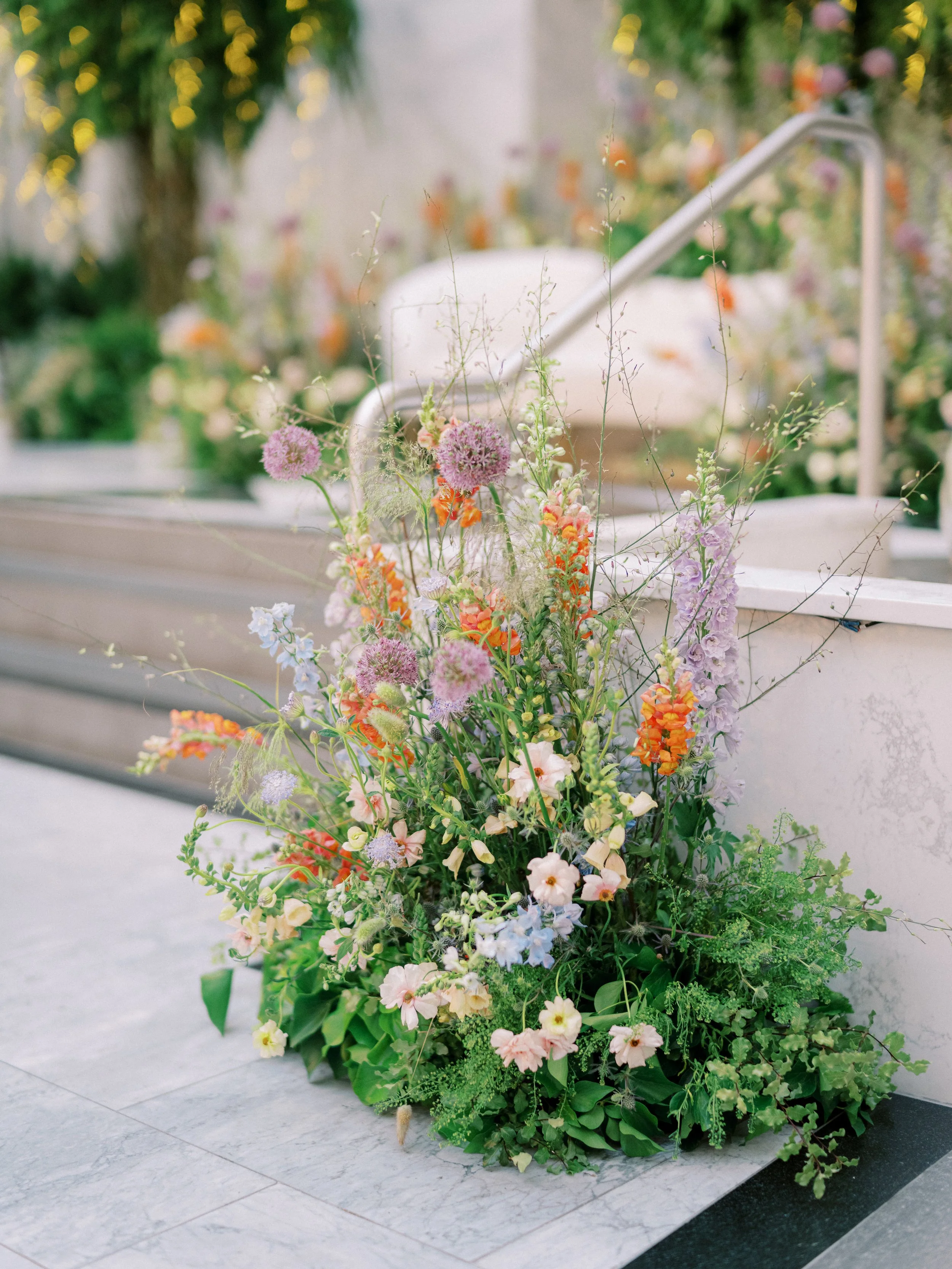 A large bouquet of colorful wildflowers arranged on a white marble surface. The bouquet includes pink, purple, orange, and white flowers with green foliage and is set outdoors with a blurred background of greenery and lights.