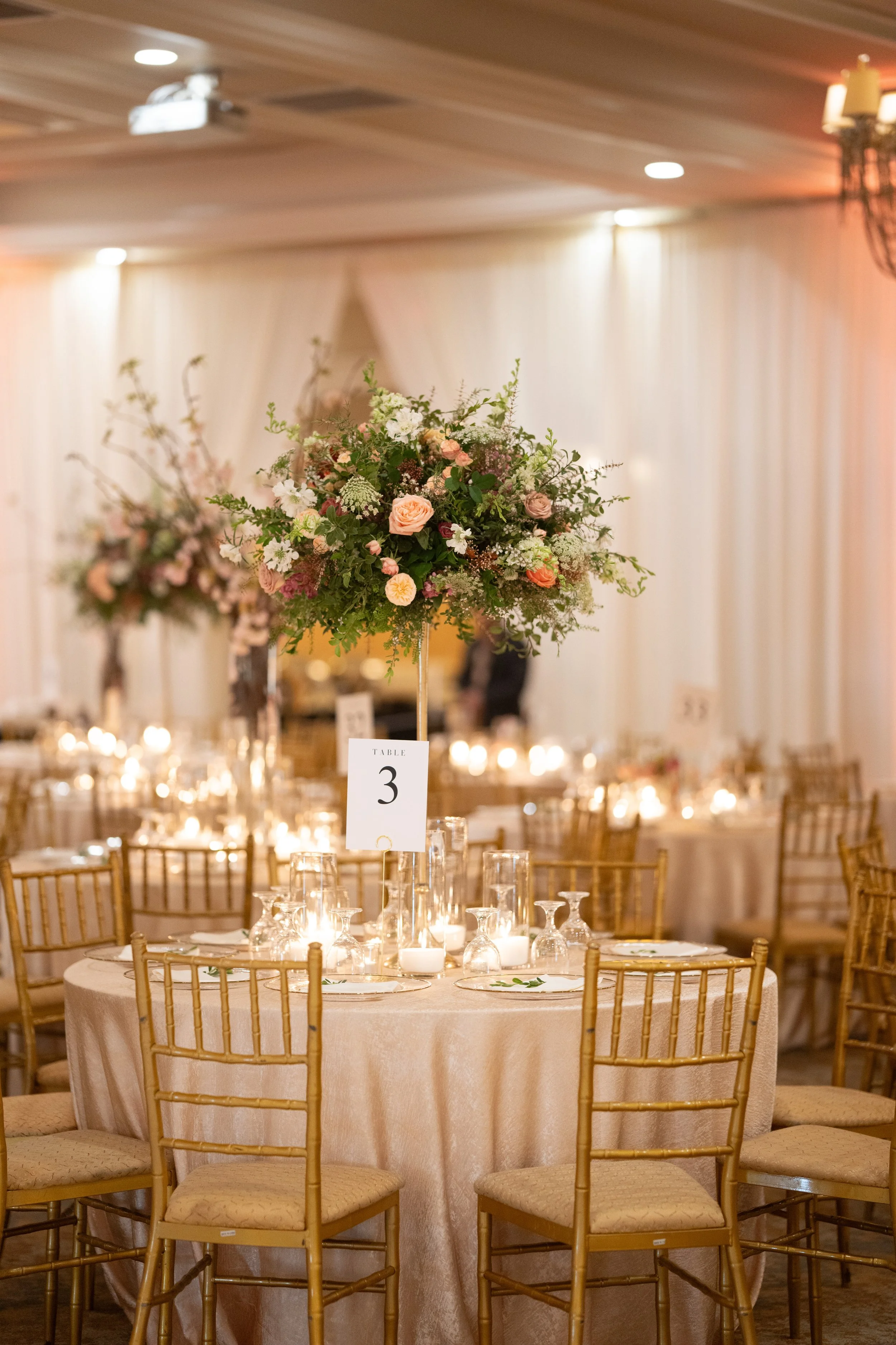 A decorated banquet table with a floral centerpiece, surrounded by gold chairs, and lit by candles, in an elegant event hall with draped curtains and overhead lighting.