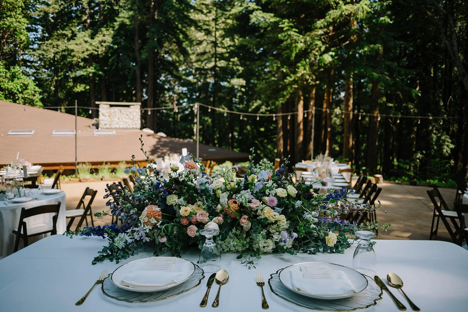 A banquet table decorated with a large floral centerpiece at an outdoor event in a wooded area. The table is set with white plates, gold utensils, and upside-down glasses, with additional tables and chairs visible in the background.