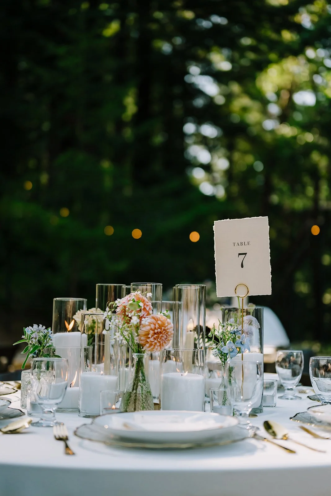 Elegant outdoor table set with candles, glassware, pink and white flowers, and a table number sign reading 'Table 7' amidst a lush green background.