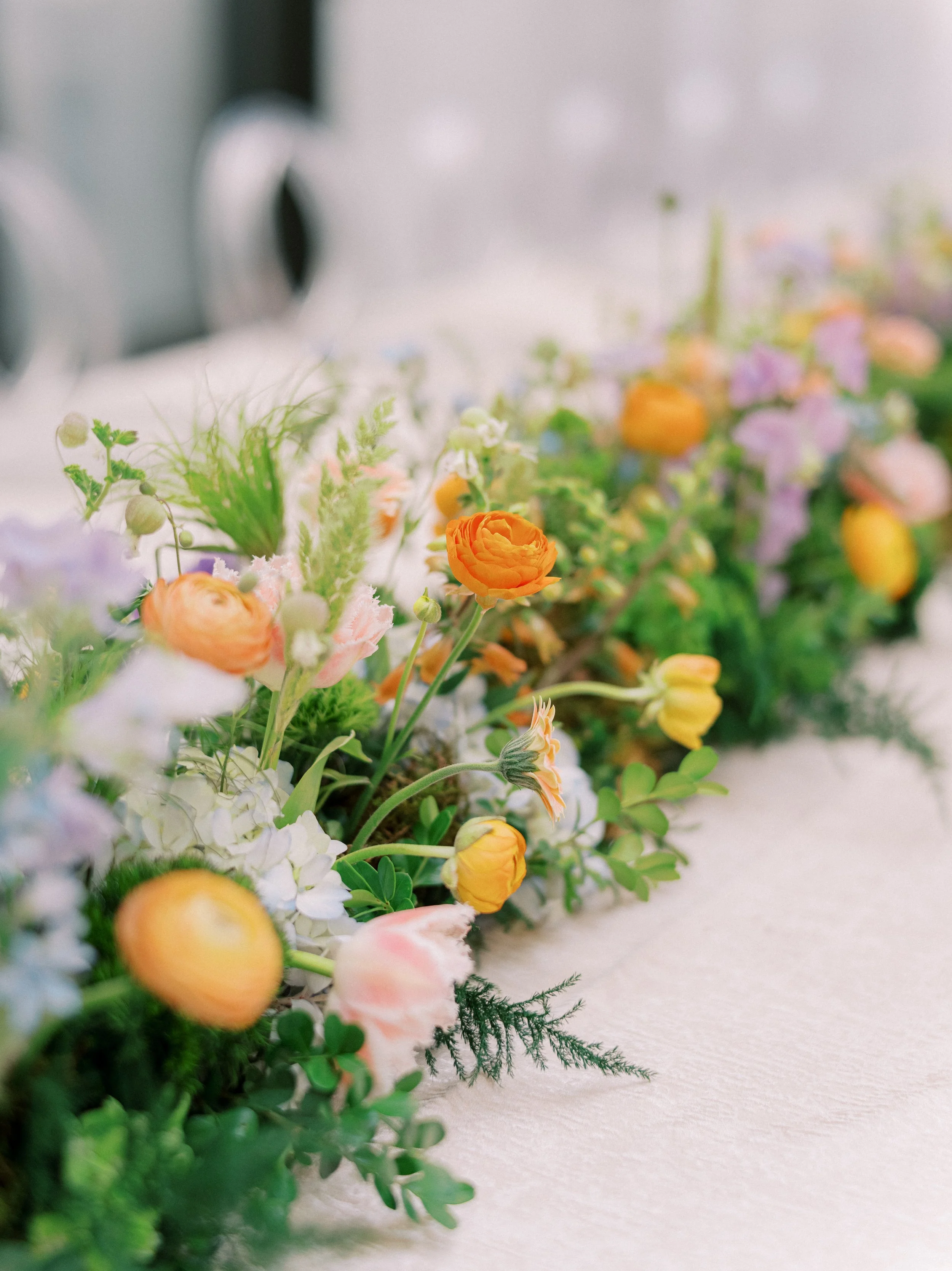 Close-up of a colorful flower arrangement with orange, pink, purple, and white flowers and greenery, placed on a white surface.