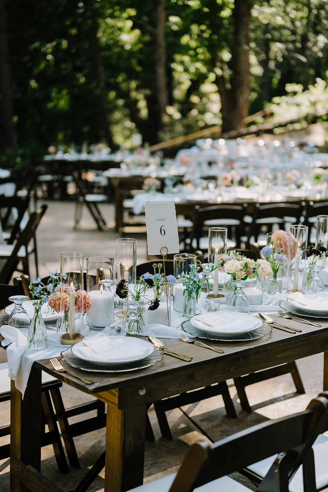 Elegant outdoor wedding reception table decorated with floral arrangements, candles, and tableware in a wooded setting.