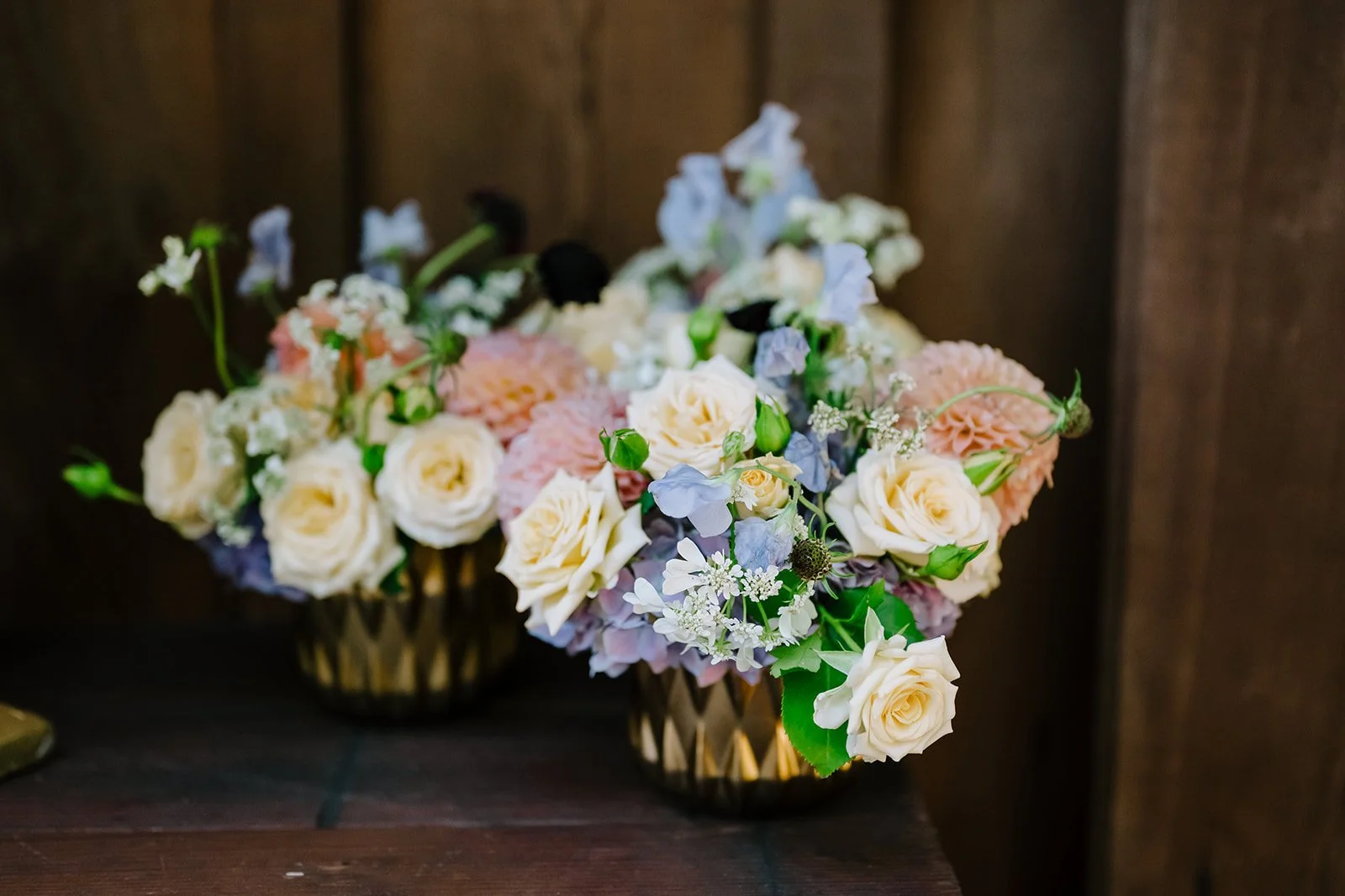 Two vases filled with a variety of fresh flowers including white roses, pink dahlias, purple delphiniums, and small white filler flowers, placed on a dark wooden surface against a wooden wall.