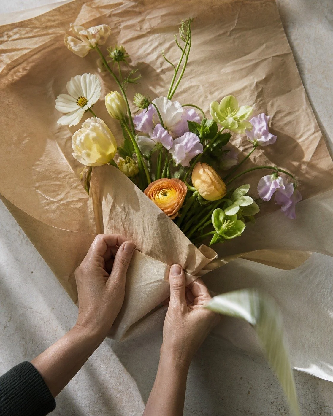 Hands wrapping a loose seasonal bouquet of ranunculus and cosmos in kraft paper