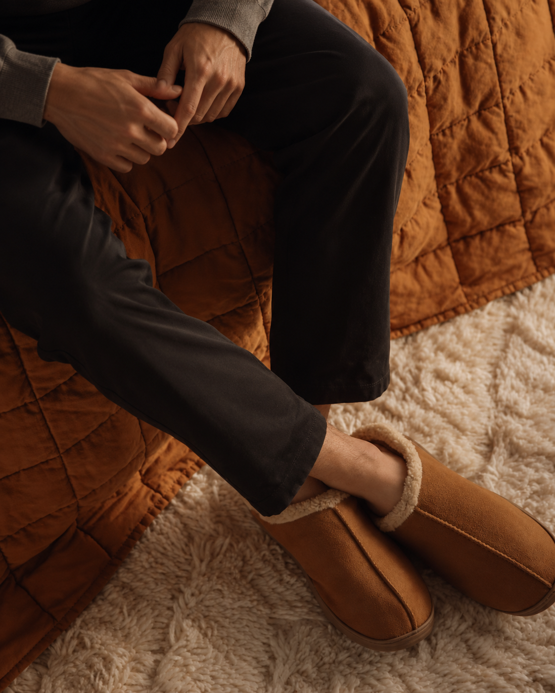 Person sitting on a quilted orange bedspread, wearing black pants and tan slippers with fluffy lining, sitting on a cream-colored shaggy rug.