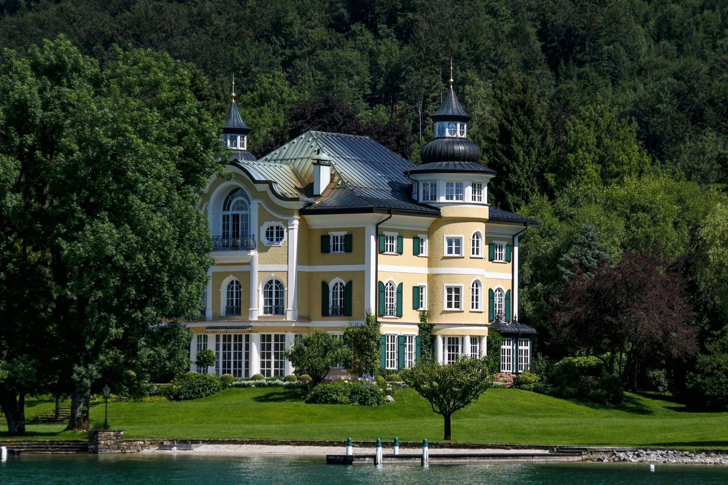 A yellow and white mansion with green shutters, large windows, and a distinctive turret with a copper roof, surrounded by lush green trees and shrubs, viewed from across a body of water.
