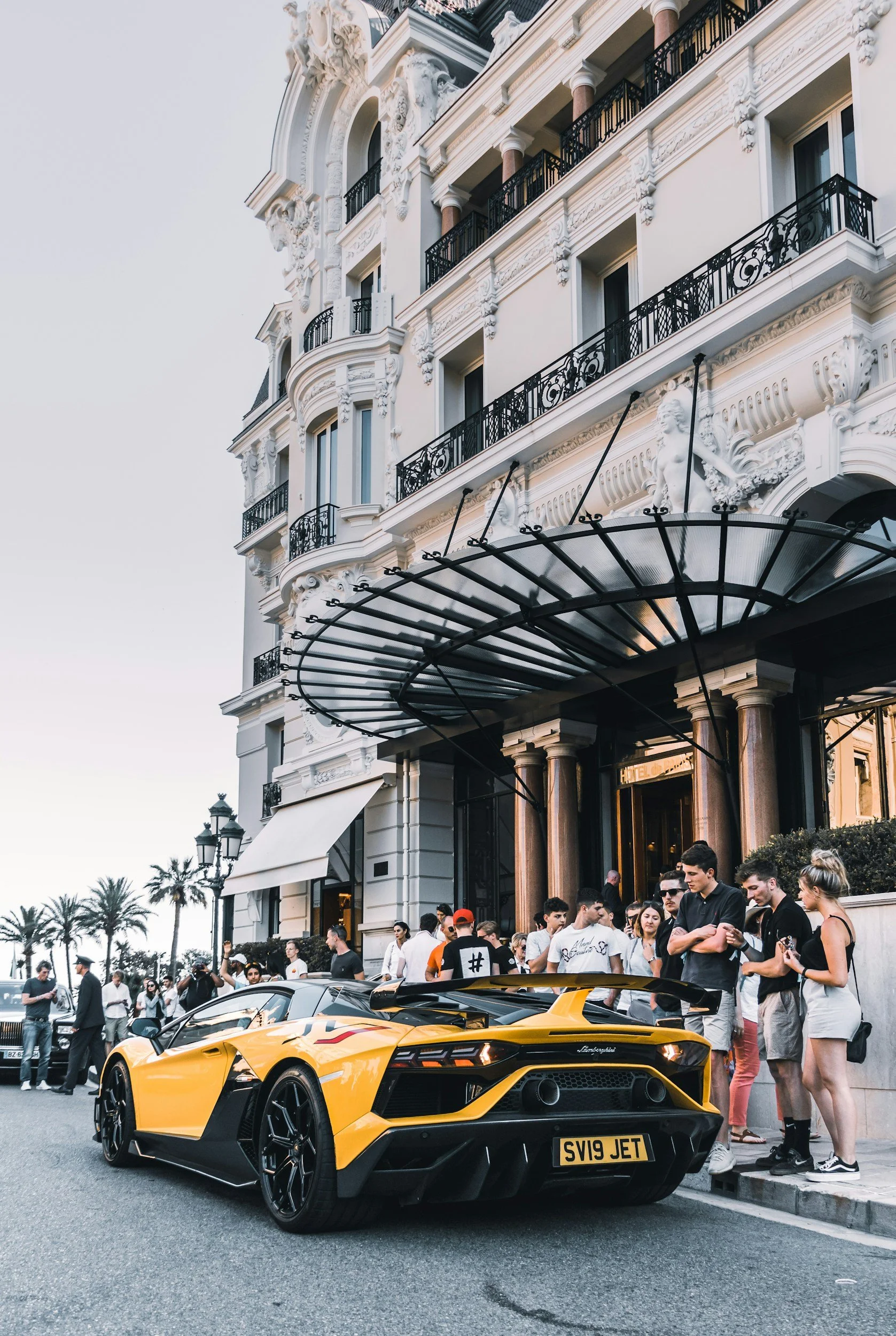 A yellow Lamborghini sports car parked on the street in front of a building with ornate architecture and a crowd of people gathered around.