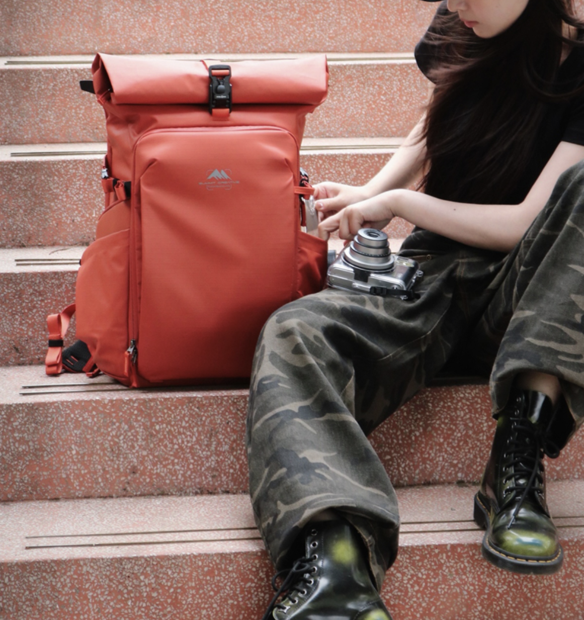 Junge Person sitzt auf roten Treppen, umgeben von einem großen roten Rucksack, und überprüft den Inhalt eines Seitentaschenreißverschlusses, während eine Kamera auf den Knien liegt.