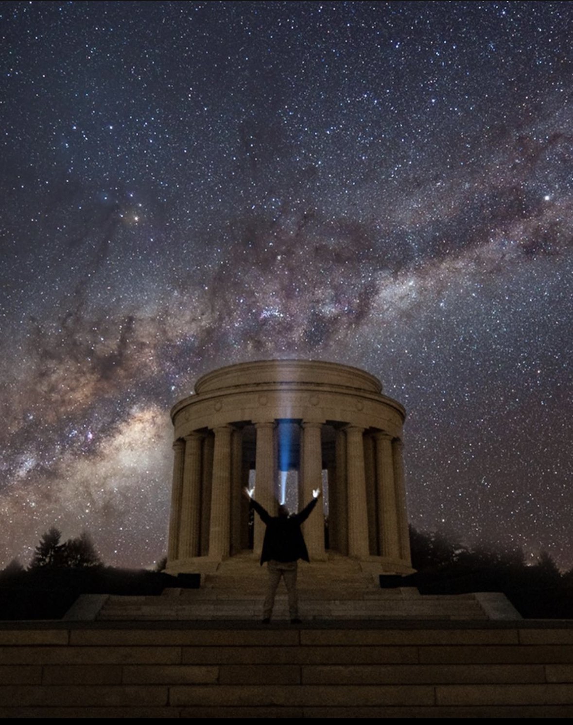 Ein Mann steht vor einem Denkmal das aus vielen Säulen besteht. Es ist Nacht und er streckt die Arme gegen den Himmel in welchem die Milchstraße zu erkennen ist.