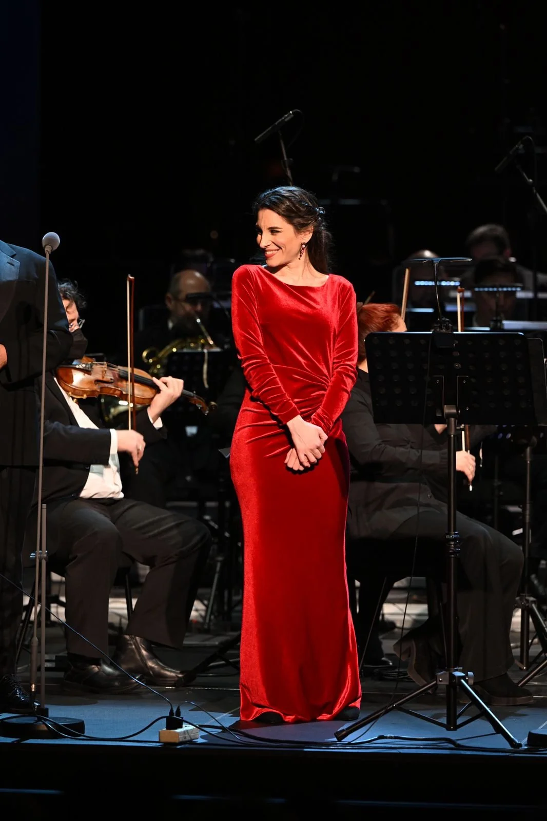 A woman in a long red dress standing on stage among an orchestra, smiling and looking to her left.