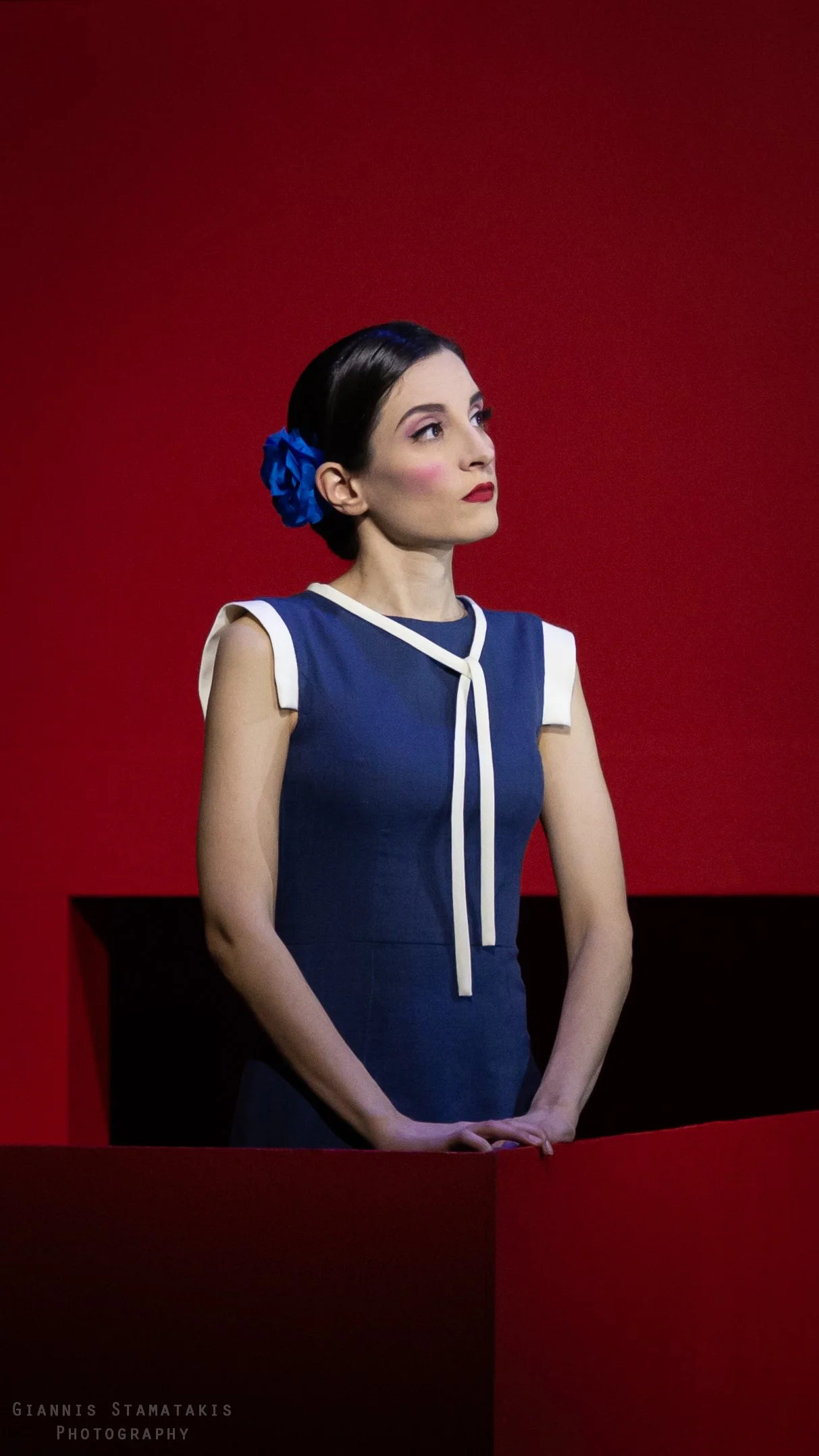 A woman with dark hair in a bun adorned with a blue flower, wearing a blue dress